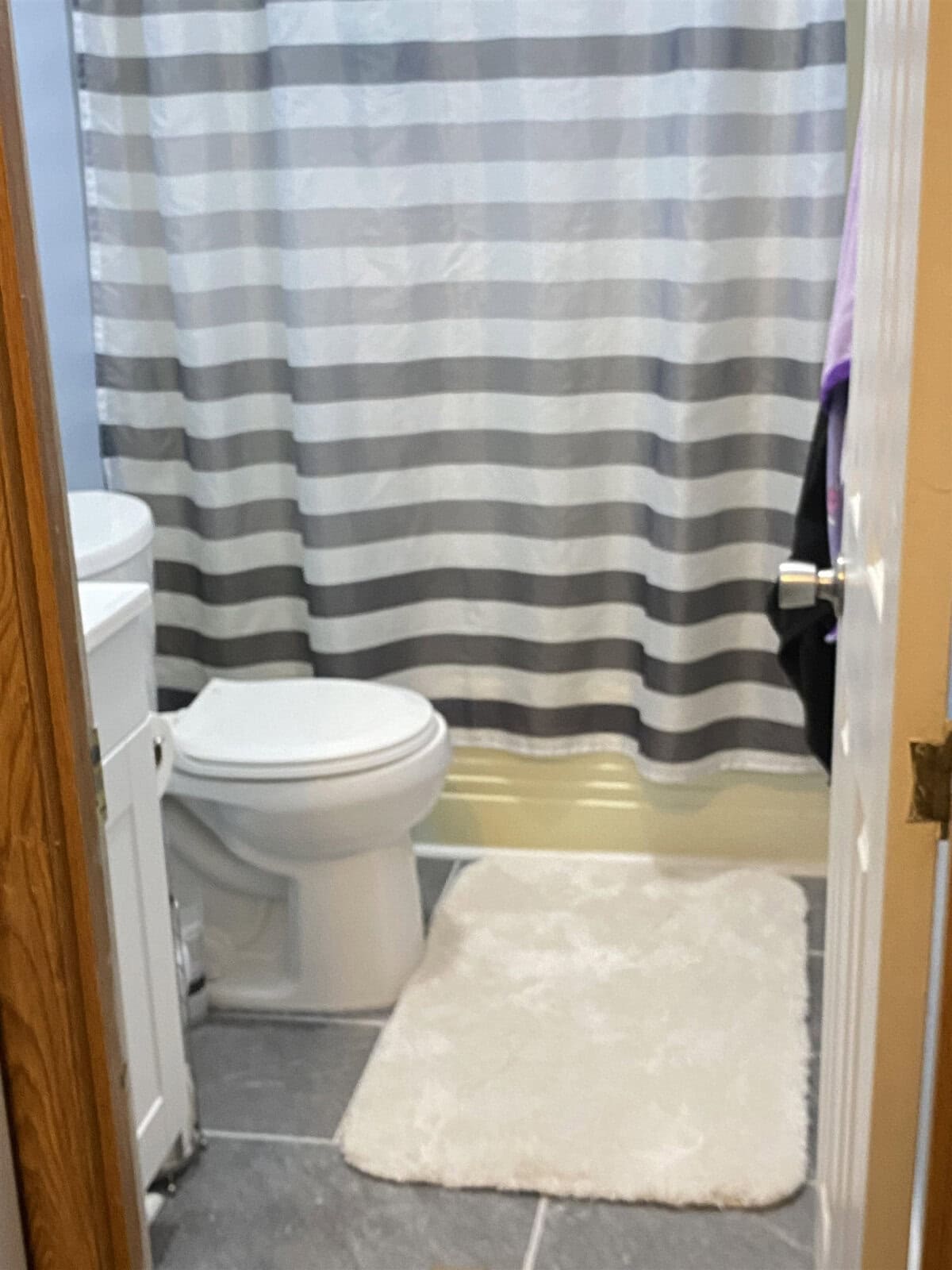 A small bathroom with a white toilet and a sink on the left. A fluffy white mat is on the gray tiled floor. The shower curtain features horizontal gray and white stripes. A door is partially open on the right.