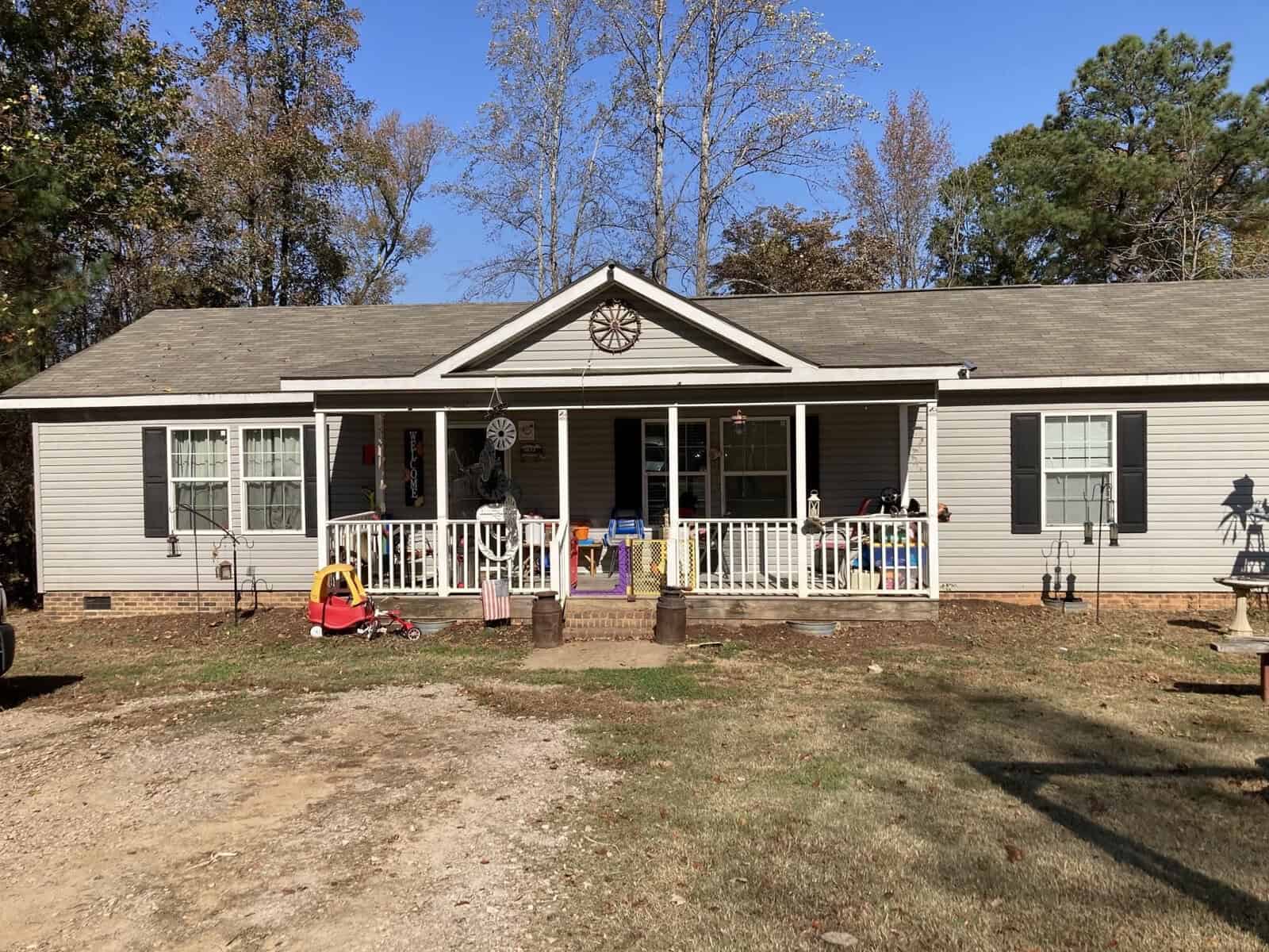 A single-story house with beige siding and a gabled porch is surrounded by trees. The porch holds various items, and a path leads from the driveway. Toys and flower pots are scattered in the yard. The sky is clear and blue.