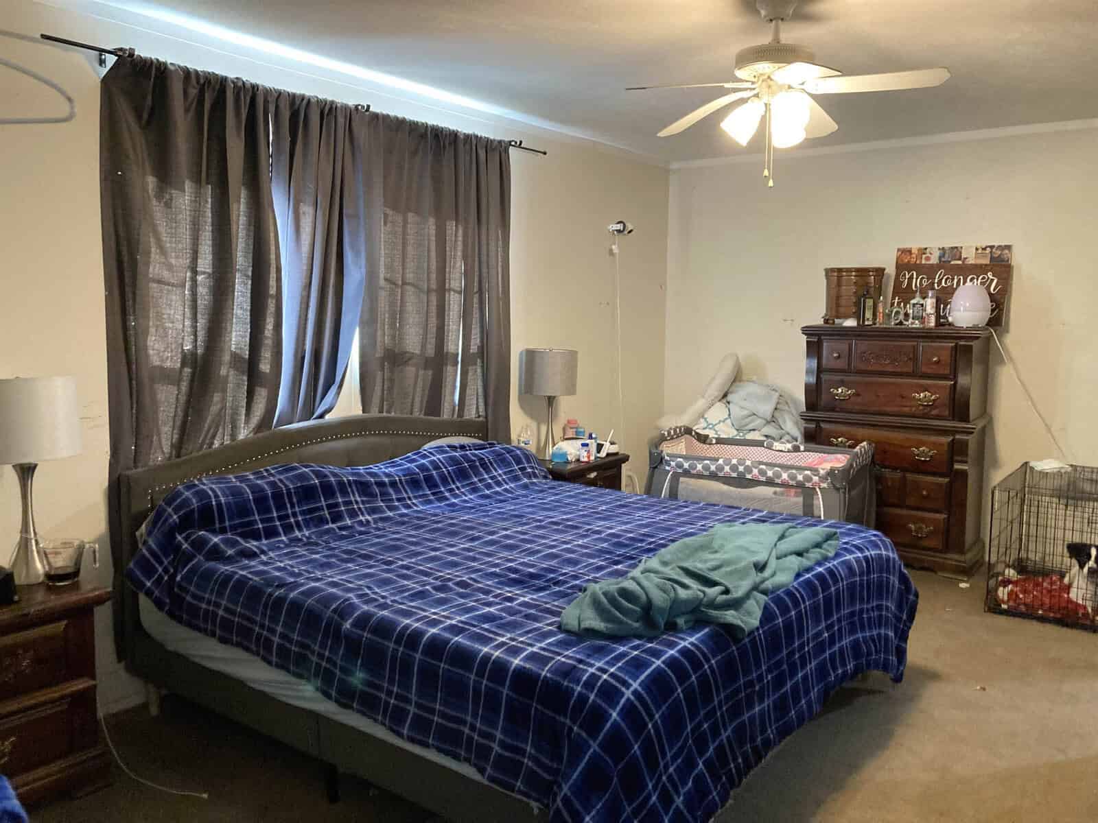 A bedroom with a large bed covered in a blue plaid blanket. There's a pile of clothes on the bed. A smaller bed is beside a wooden dresser with various items on top. A ceiling fan and thick curtains complete the room's decor.