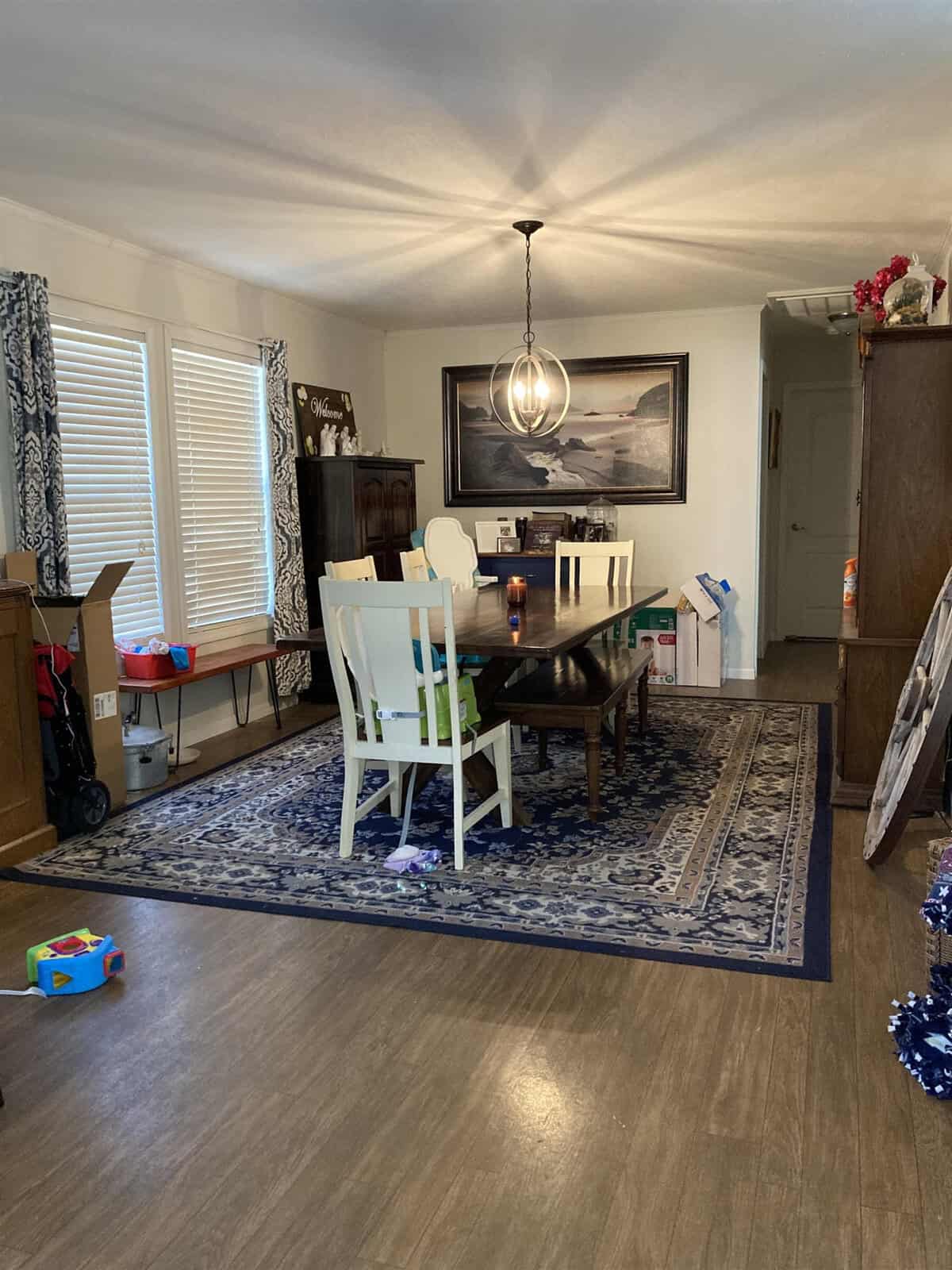 A dining room with a rectangular table, six chairs, and a decorative chandelier above. A blue-patterned rug covers part of the wooden floor. Toys are scattered and a large picture hangs on the wall. Natural light from the windows brightens the room.