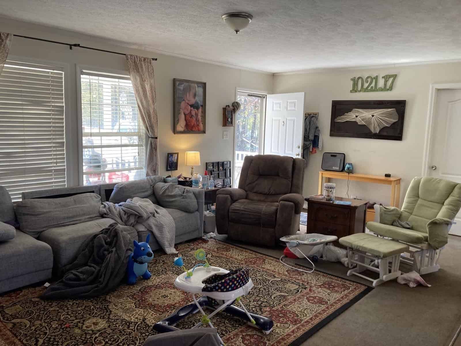 A cozy living room with a sofa, recliner, and rocking chair on a patterned rug. A baby walker and toys are scattered on the floor. Sunlight streams through the window with blinds. A painting and decor hang on the walls.