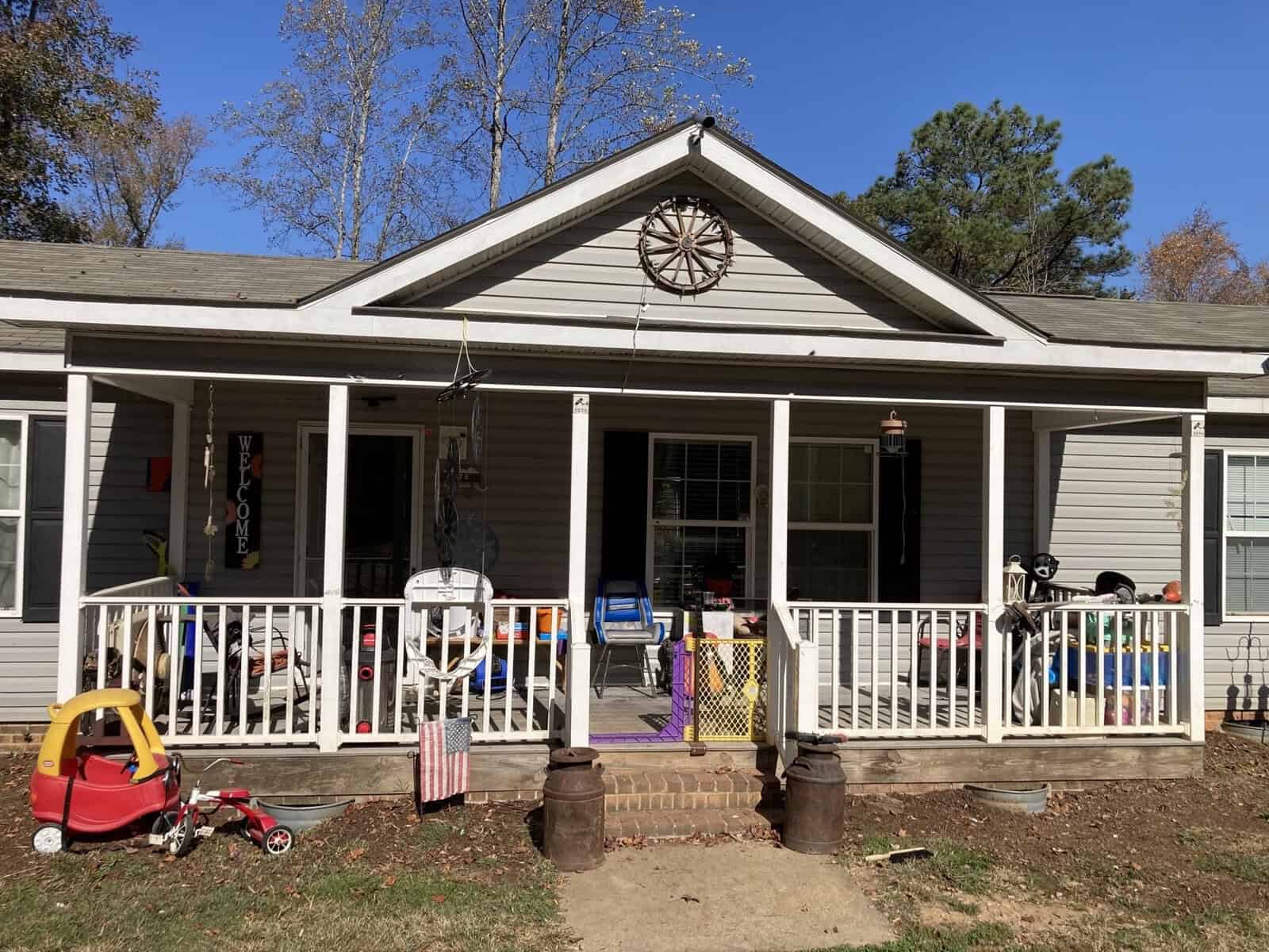 A house with a front porch filled with various items, including a child's toy car and a tricycle. The porch has a welcome sign, hanging decorations, and a wooden wagon wheel above the roof. The yard is bordered by trees under a clear blue sky.