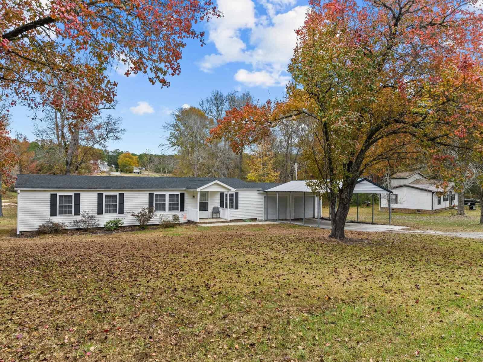 A white, single-story house with dark shutters on 2204a Sam Moss Hayes Road is surrounded by autumn trees. A carport is attached to the right side, and fallen leaves cover the grass in the spacious Creedmoor yard under a partly cloudy sky.