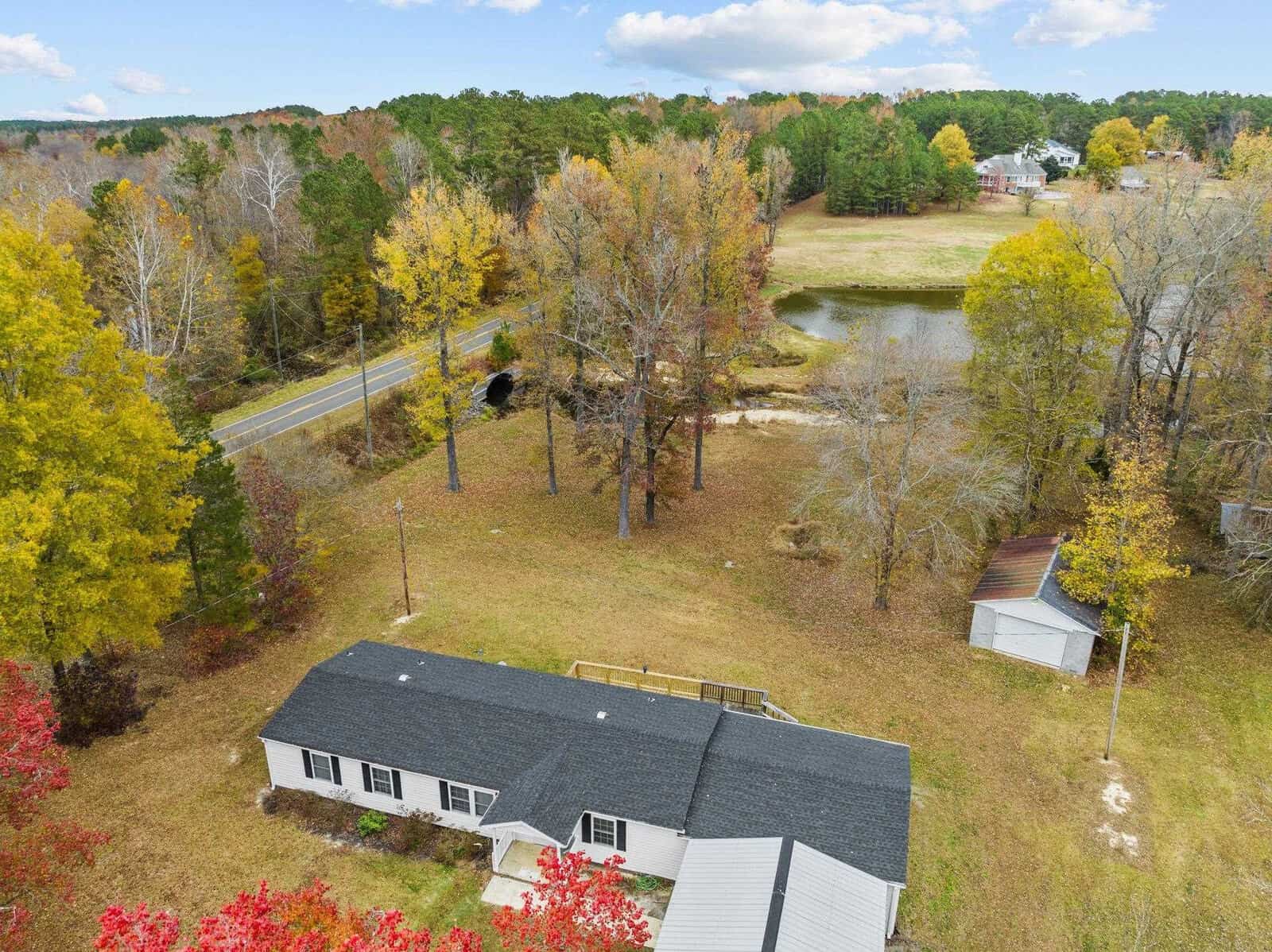 Aerial view of a rural area featuring a single-story house with a black roof, surrounded by autumn-colored trees. A small shed and a pond are nearby, with a road running alongside the property.