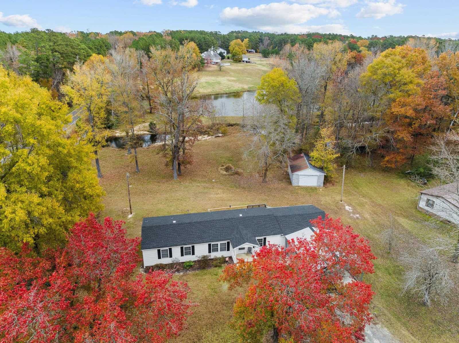 Aerial view of a house surrounded by vibrant autumn trees in red and orange hues. A small pond is visible in the background, and a white shed is near the house. The sky is partly cloudy, and the landscape is lush and serene.