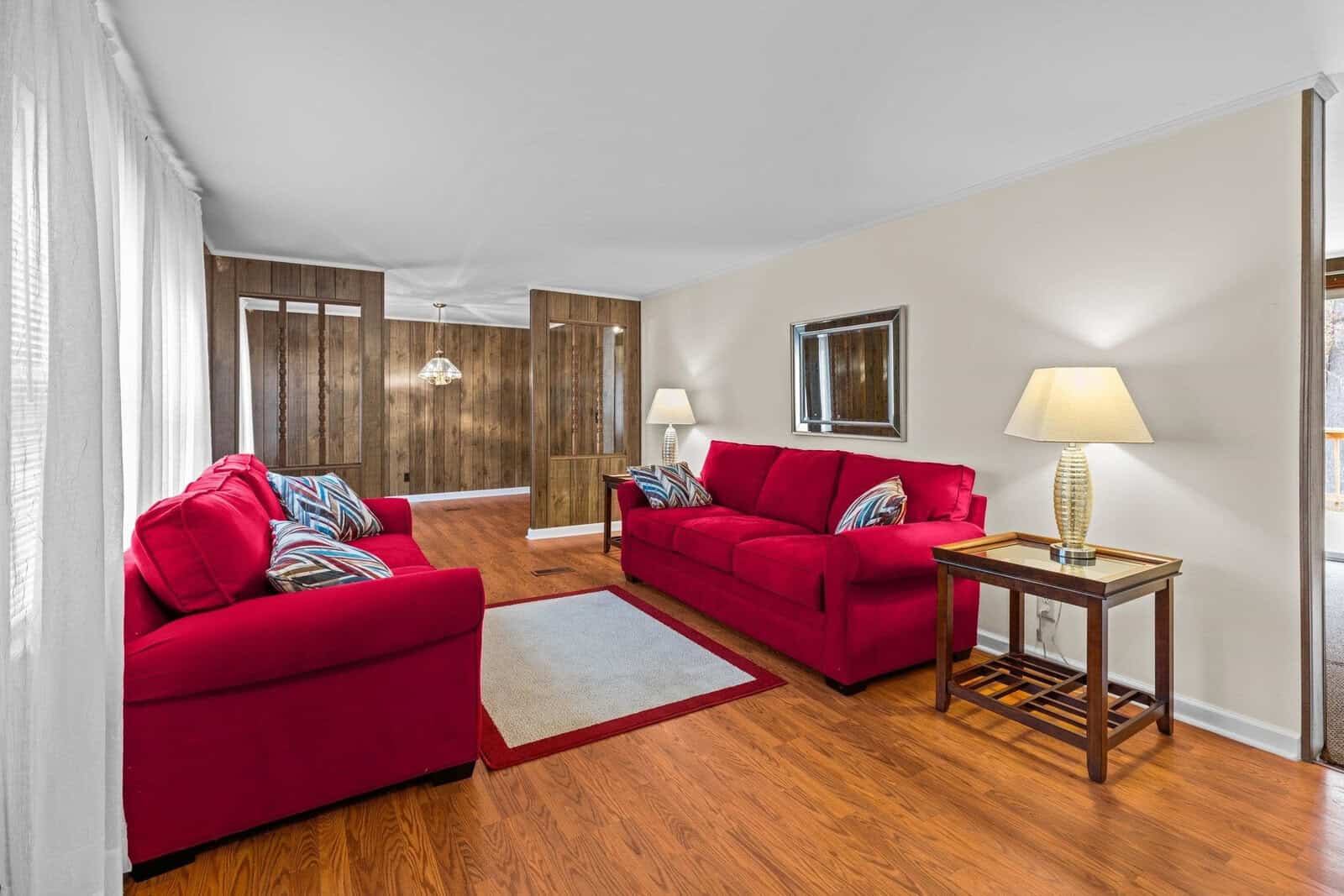 A cozy living room with wooden floors, featuring two red sofas with patterned pillows. There's a rectangular rug between them, two side tables with lamps, a wall mirror, and a view into a dining area with dark wooden walls and a hanging light fixture.