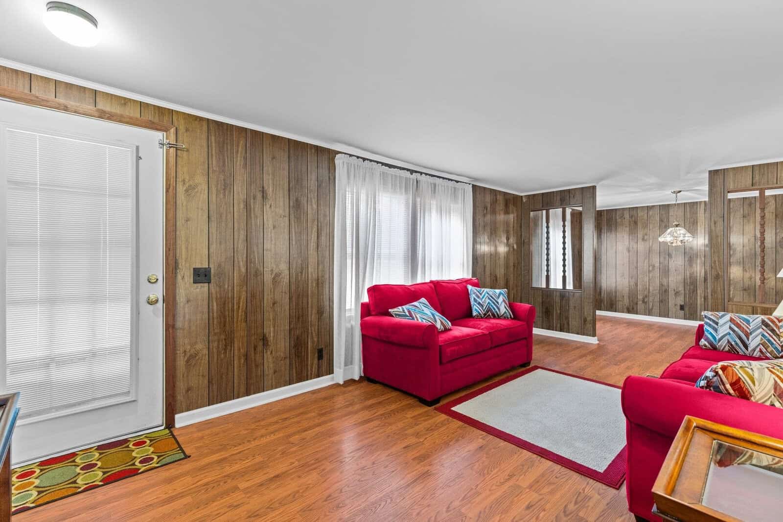 A cozy living room with wood-paneled walls and hardwood floors. Red sofas with patterned throw pillows face each other, situated on a light gray rug. A glass light fixture hangs in the background, and a white door is on the left.