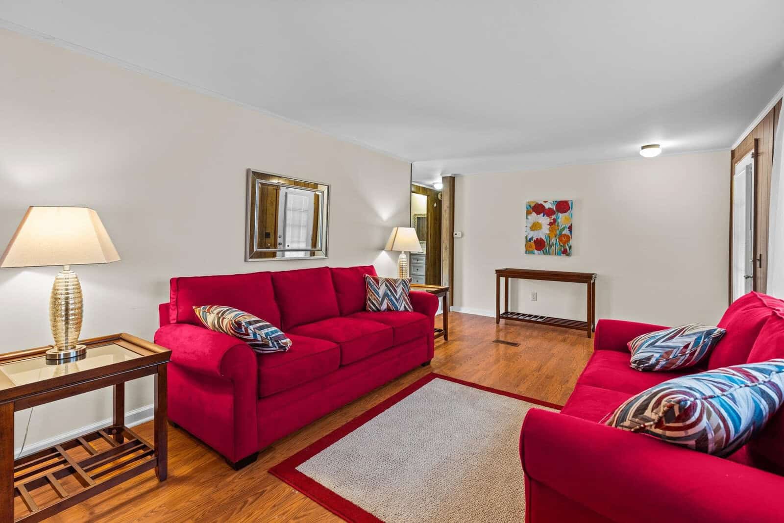 A cozy living room with two vibrant red sofas, accented with patterned throw pillows. A neutral rug lies on the wooden floor. A side table with a lamp is beside one sofa. Artwork and a mirror adorn the beige walls, enhancing the room's warmth.