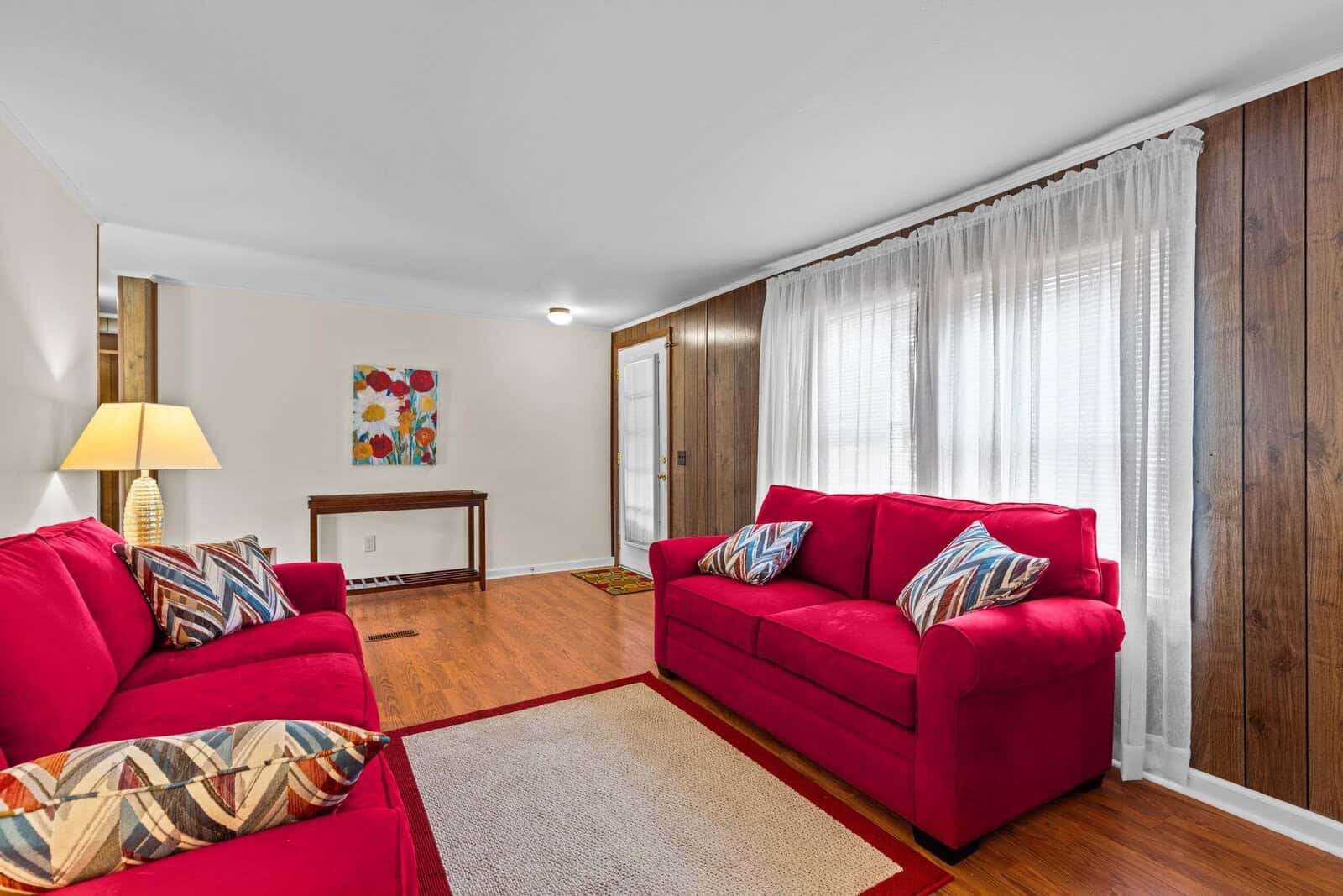 A cozy living room with two red sofas, colorful patterned pillows, and a lamp on the side. A small table against the wall holds a bright floral painting. The room has wood-paneled walls and a large window with sheer curtains allowing natural light.