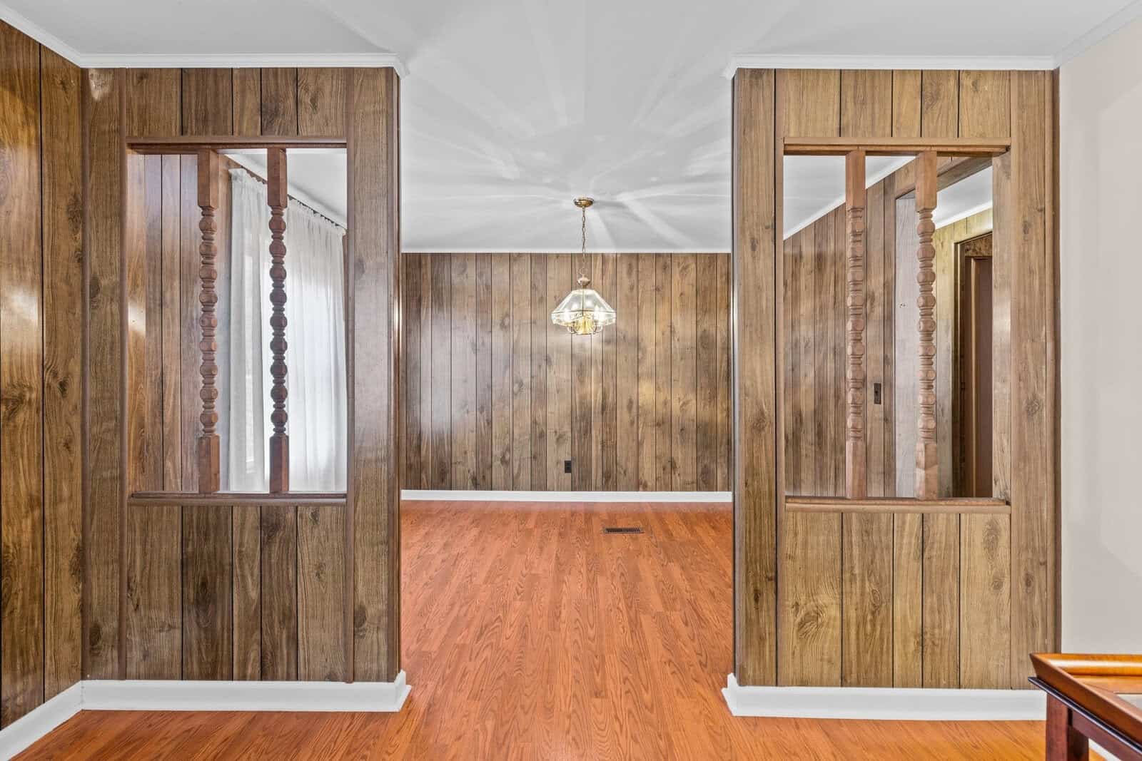 Interior view of a room with wood-paneled walls and wood flooring. Decorative wooden partitions divide the space. A hanging light fixture is centered in the far room, and a window with sheer curtains is visible on the left.