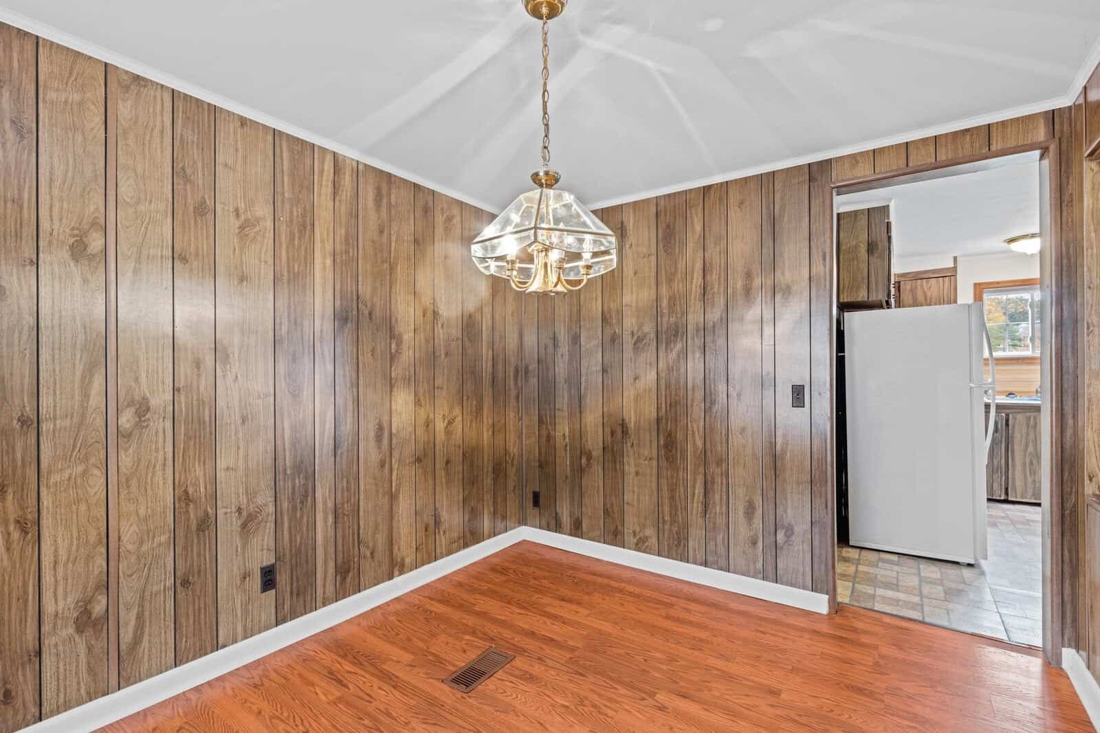 A room with wood-paneled walls, a hardwood floor, and a geometric glass pendant light. An open doorway leads to a kitchen with visible flooring and a refrigerator. The space is empty and well-lit.