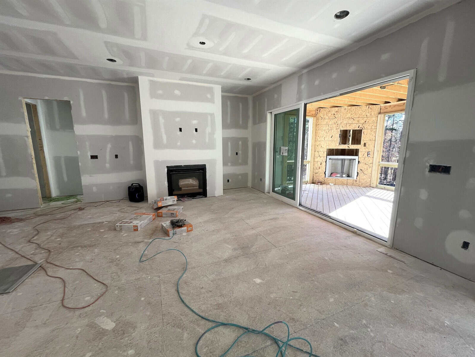 A room under construction with unpainted drywall, a fireplace, and large sliding glass doors leading to a porch. Boxes and cables are scattered on the floor. The outdoor porch also has a fireplace and wooden walls.