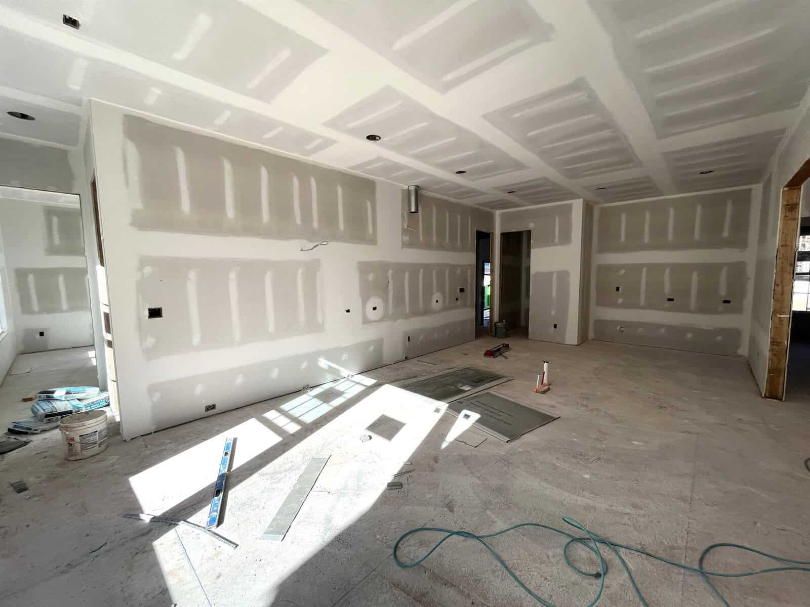 A partially constructed room with unfinished drywall on the walls and ceiling. Construction materials, including a can of paint and tools, are scattered on the floor. A doorway is visible, and sunlight streams in through a window on the right.