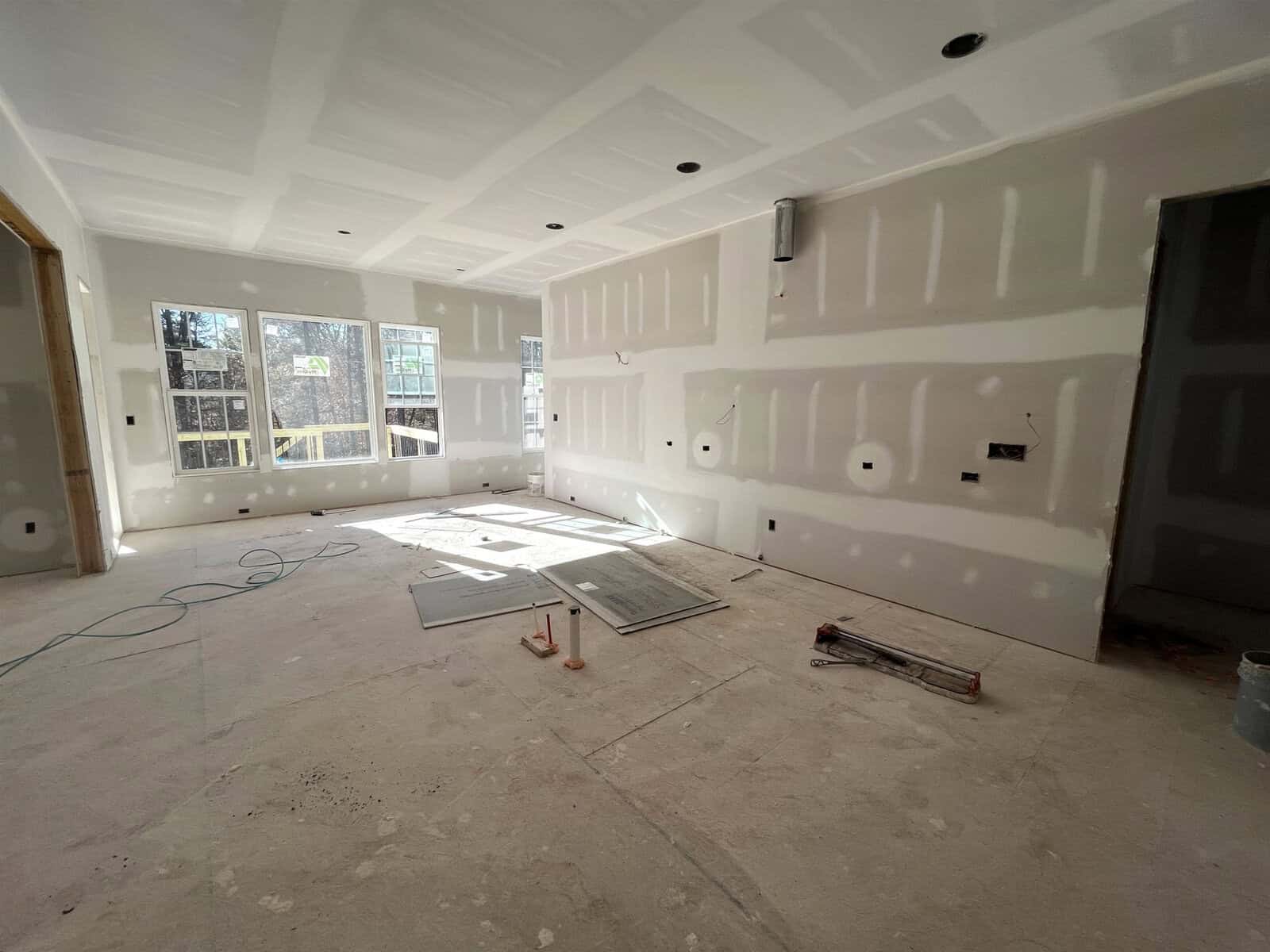 A room under construction with unfinished drywall on the walls and ceiling. The floor is covered with construction materials, including panels and tools. Sunlight streams in through three large windows. Wires and light fixtures are visible.