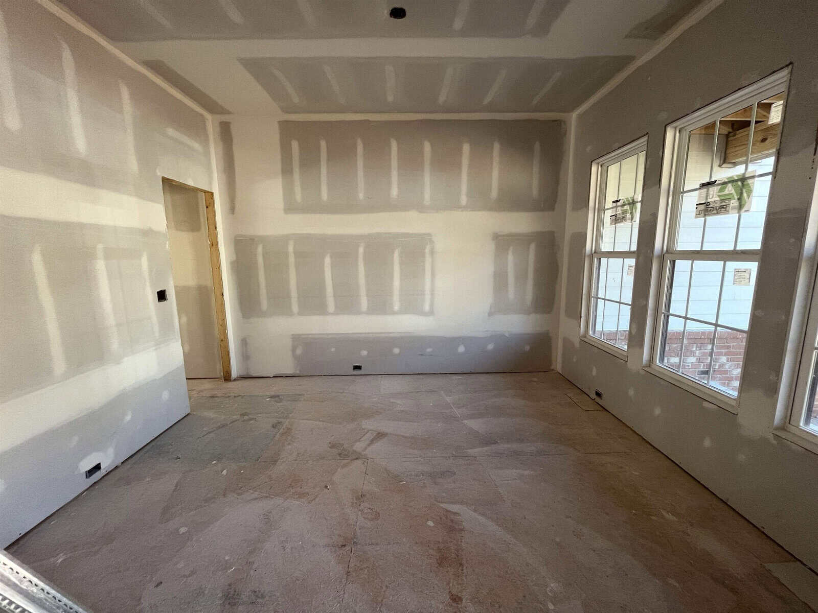 A room under construction with drywall installed on the walls and ceiling. The floor appears unfinished, and there are two large windows on one wall. A door opening is visible on the left. The room is empty.