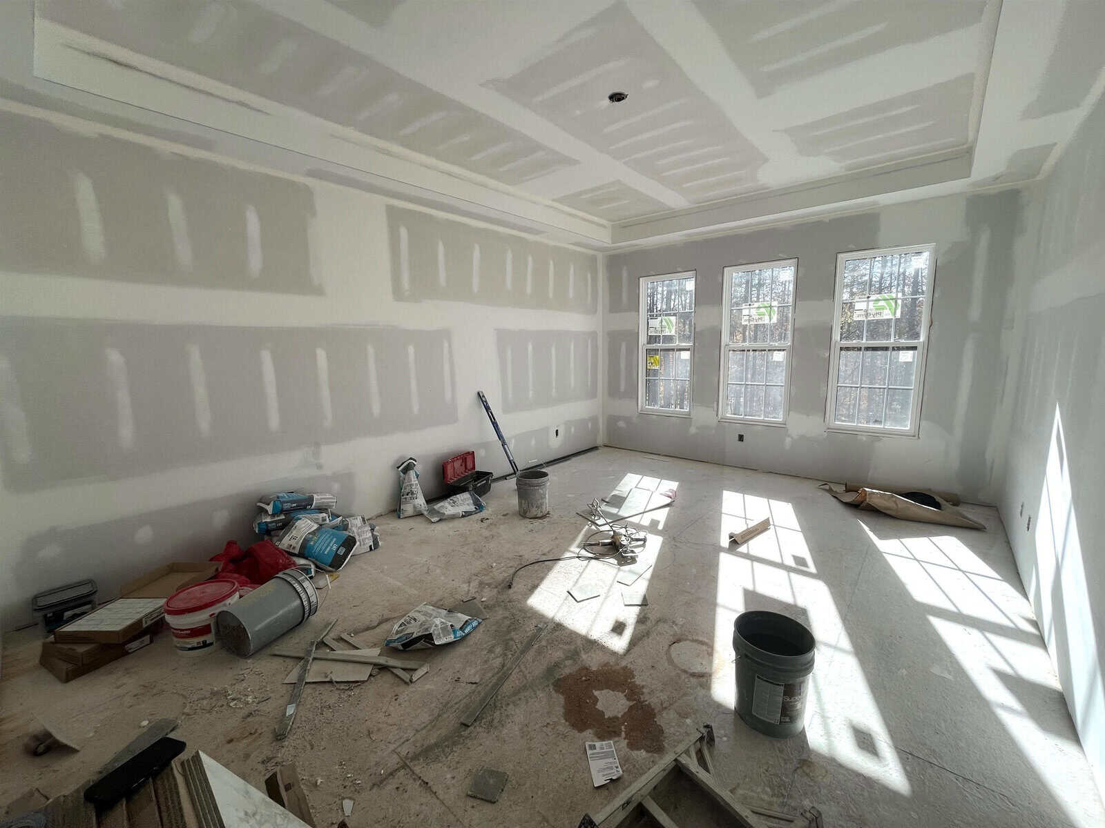 A room under construction with unfinished drywall. The floor is scattered with tools, buckets, and materials. Sunlight streams through three large windows at the far end, casting shadows across the floor.
