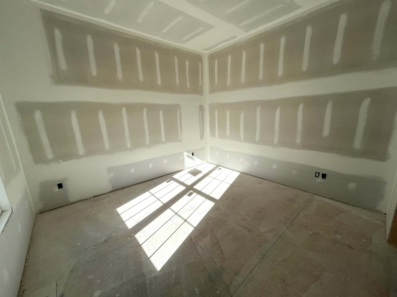 An empty room with unfinished drywall and visible joints. Sunlight enters through a window, creating a grid pattern on the floor. The room has no flooring or furnishings, with electrical outlets visible on the walls.