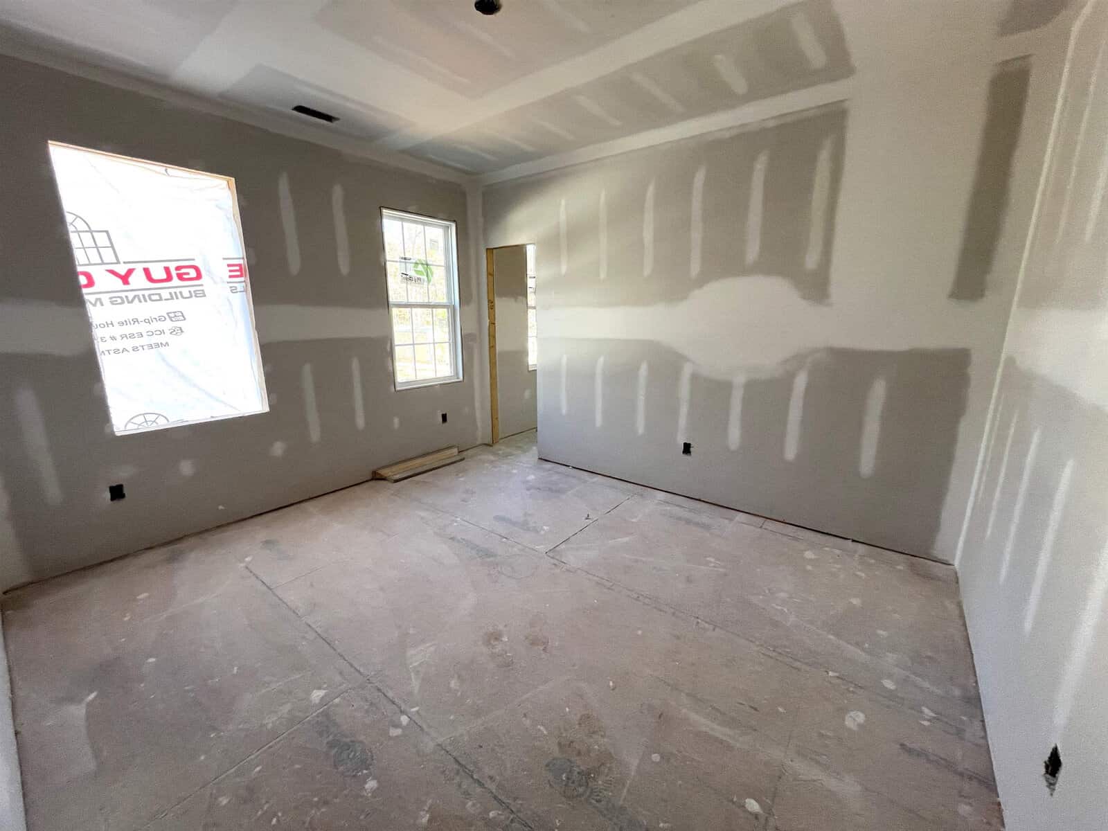 An unfinished room with drywall installation in progress. The walls and ceiling have visible seams and fasteners. There are two windows on one wall, letting in natural light. The floor is bare, revealing a rough surface. No furniture is present.