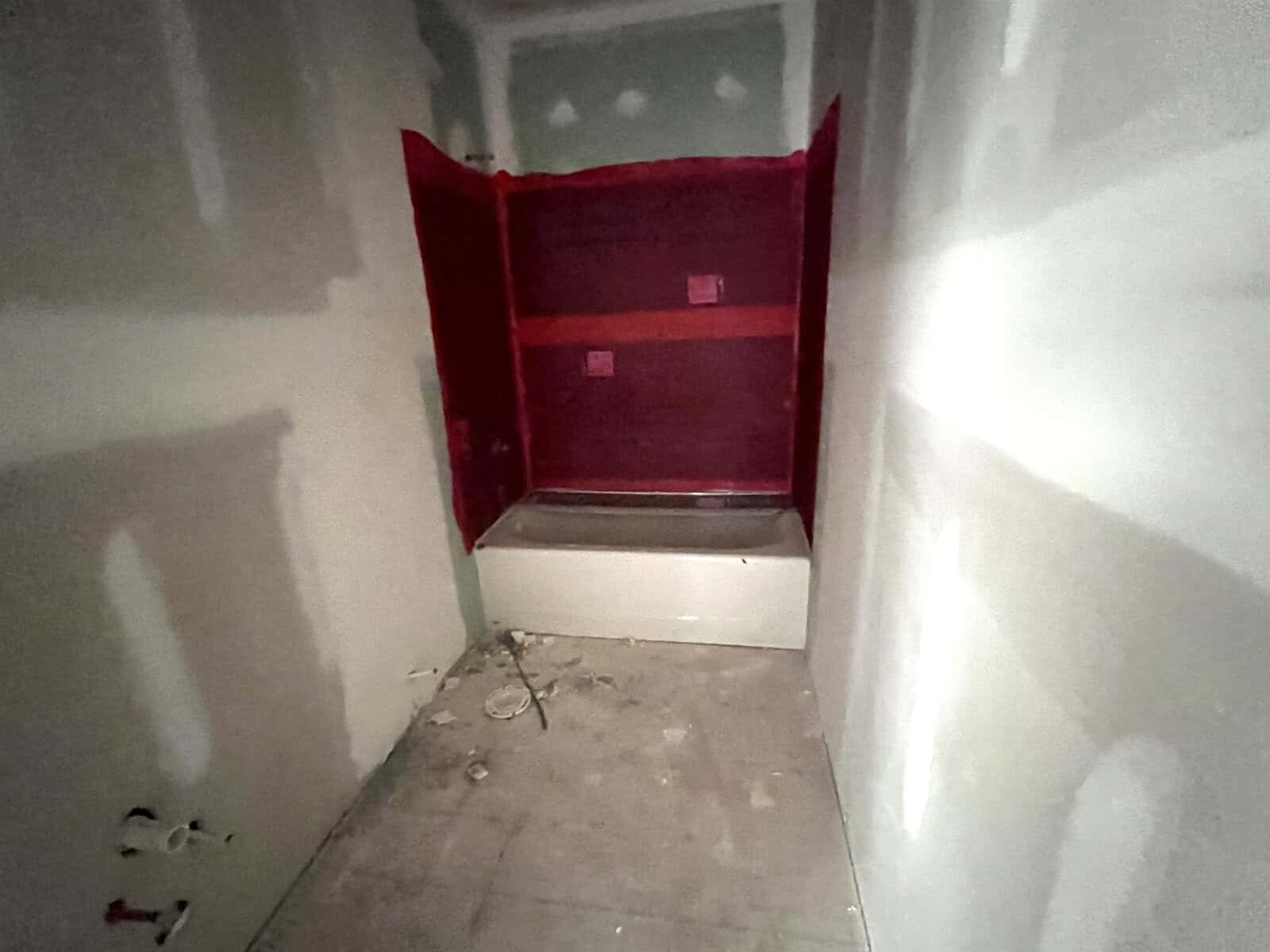 A dimly lit, unfinished bathroom with unpainted drywall. A red waterproof membrane covers the shower area above a white bathtub. Plumbing fixtures are visible on the left, and the concrete floor is bare with some construction debris.