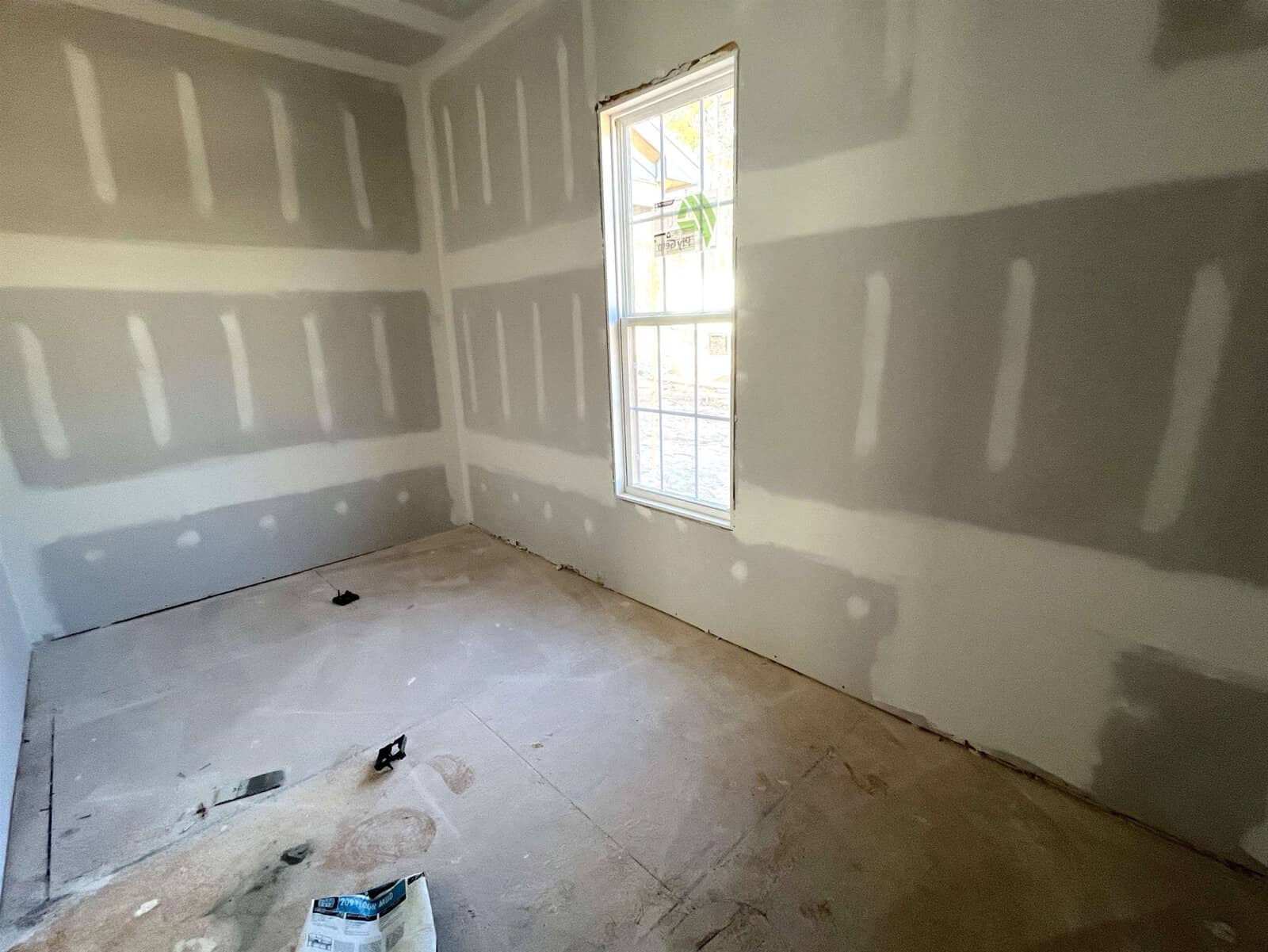 A small, unfinished room with unpainted drywall and a single window. The floor is bare, showing construction materials and debris. Light enters through the window, illuminating the grey walls with white plaster seams.