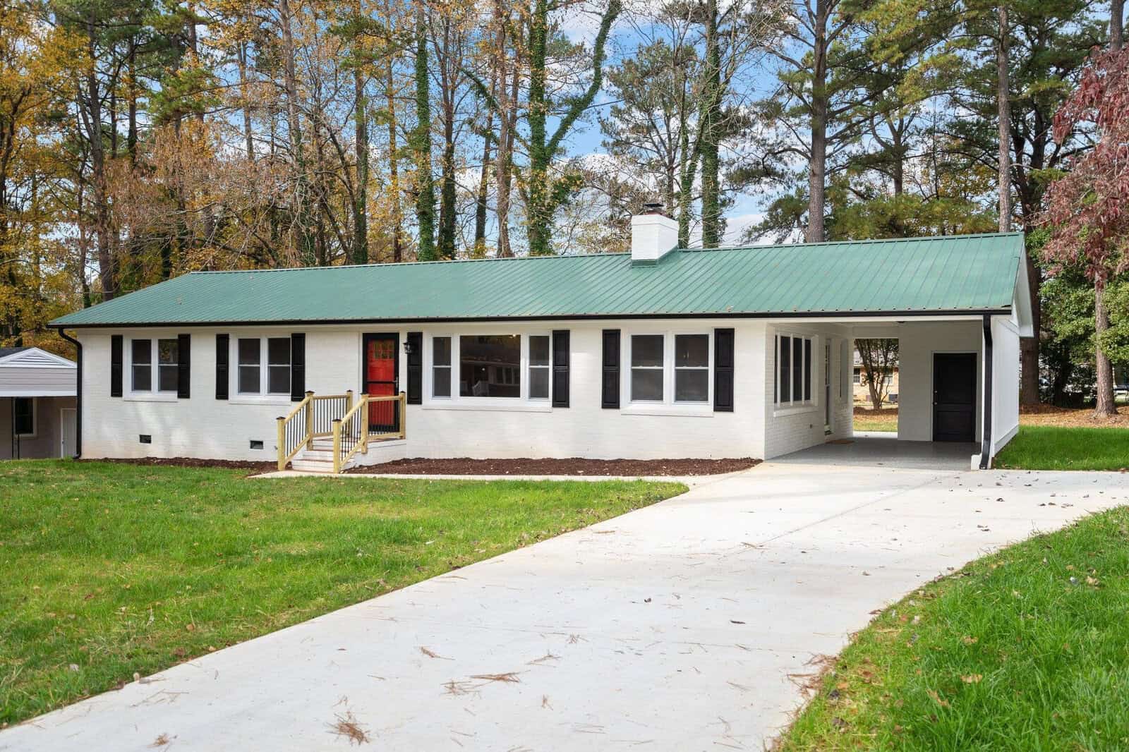A single-story residential home at 1508 Lynne Avenue, Henderson NC, with a green metal roof, white siding, and a red front door, set against a backdrop of autumn trees.