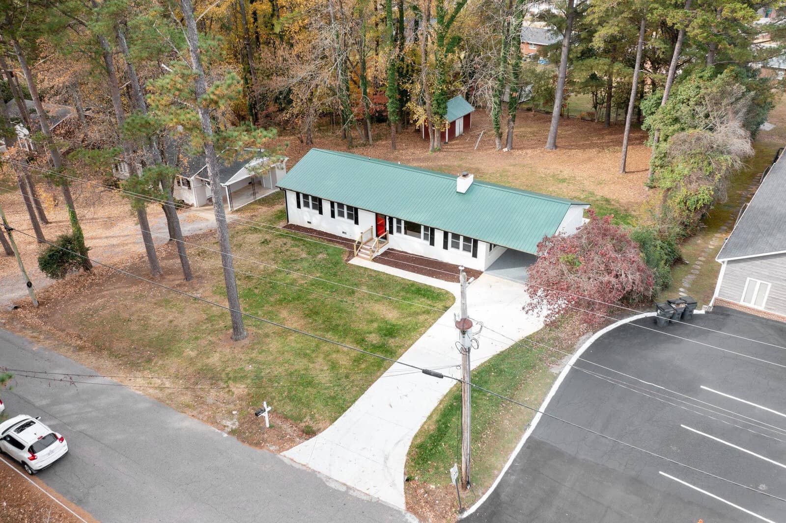 Aerial view of a single-story house with a green roof, a long driveway, and surrounding trees, adjacent to a parking area with a parked car.