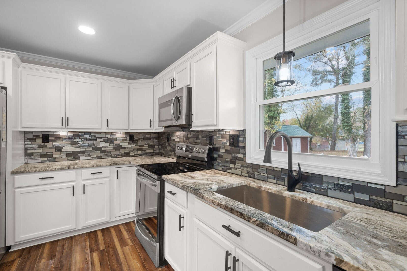 A modern kitchen with white cabinetry, stainless steel appliances, and a multi-colored backsplash, featuring hardwood floors and a view of the exterior through a window.