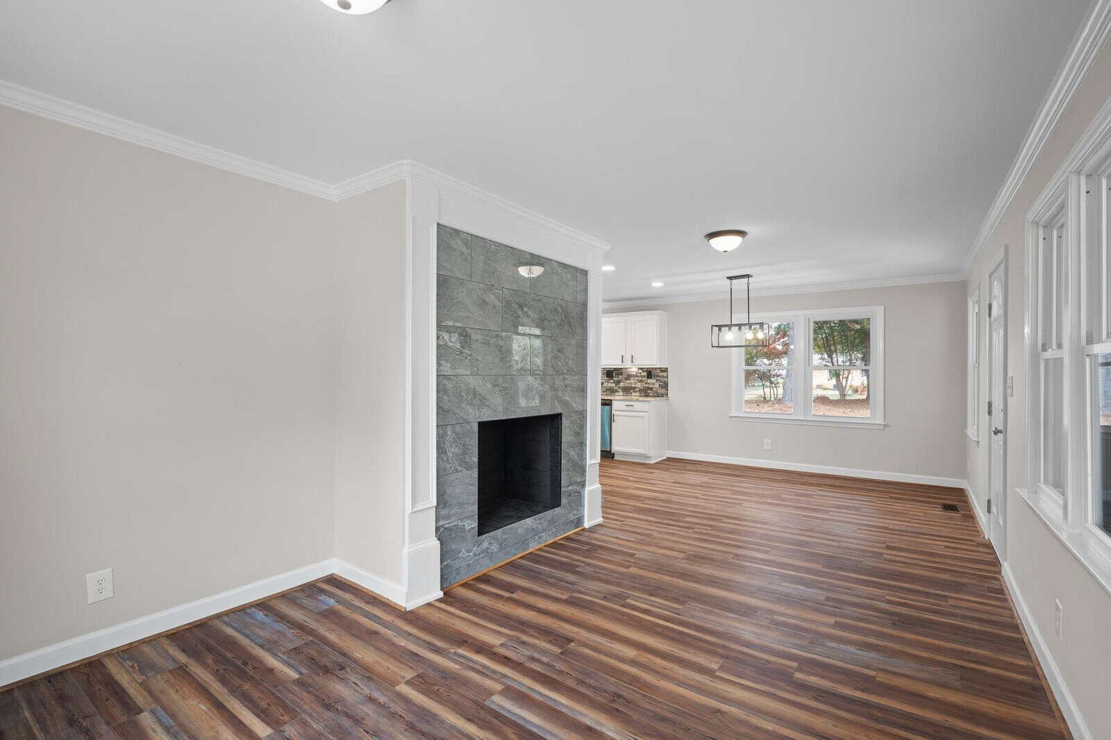 An empty living room with polished hardwood floors, white walls, a gray tiled fireplace, and a window providing natural light.