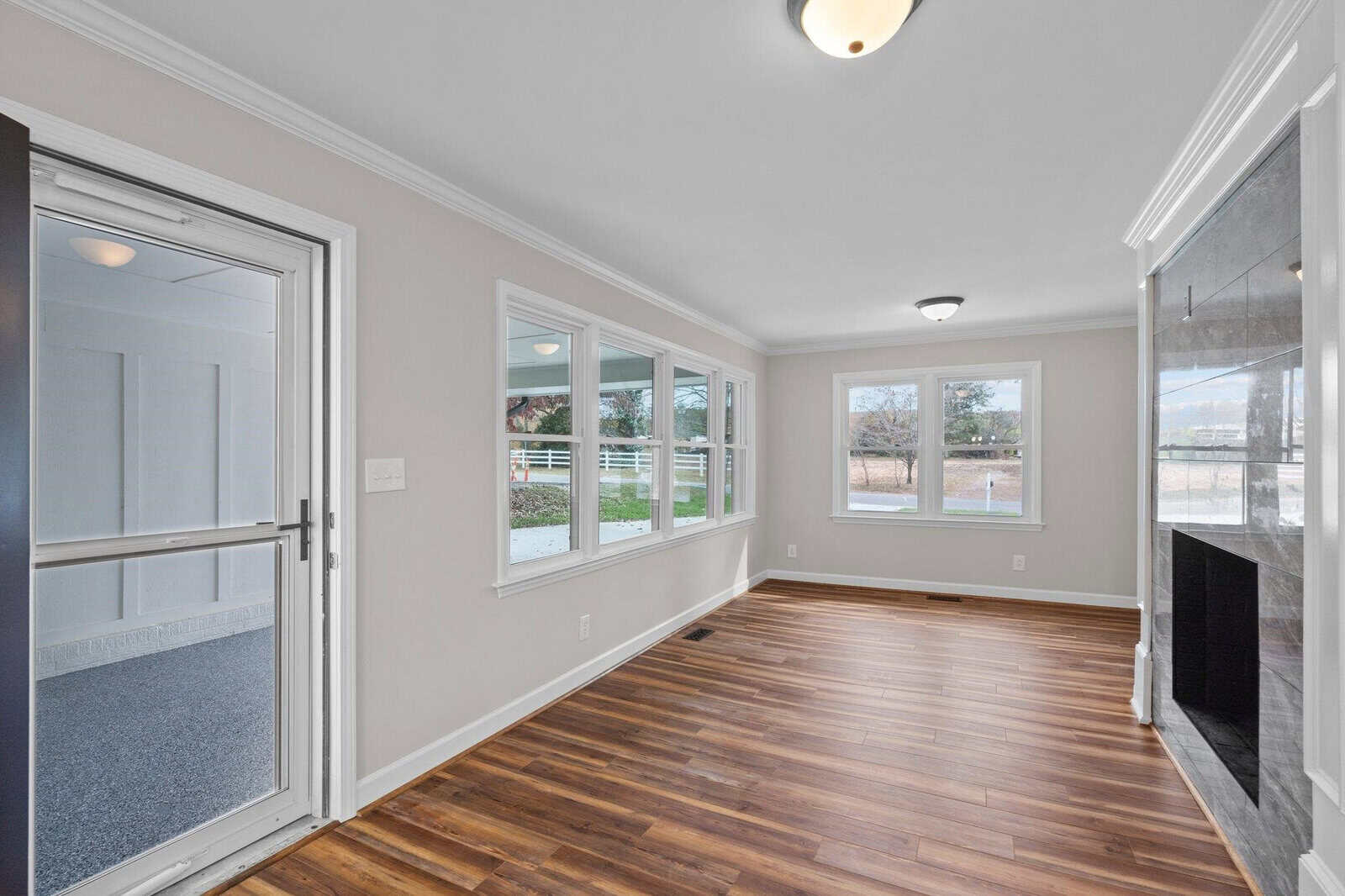 An empty living room with hardwood flooring, white walls, and multiple windows providing natural light. there is a glass door on the left and a fireplace on the right.