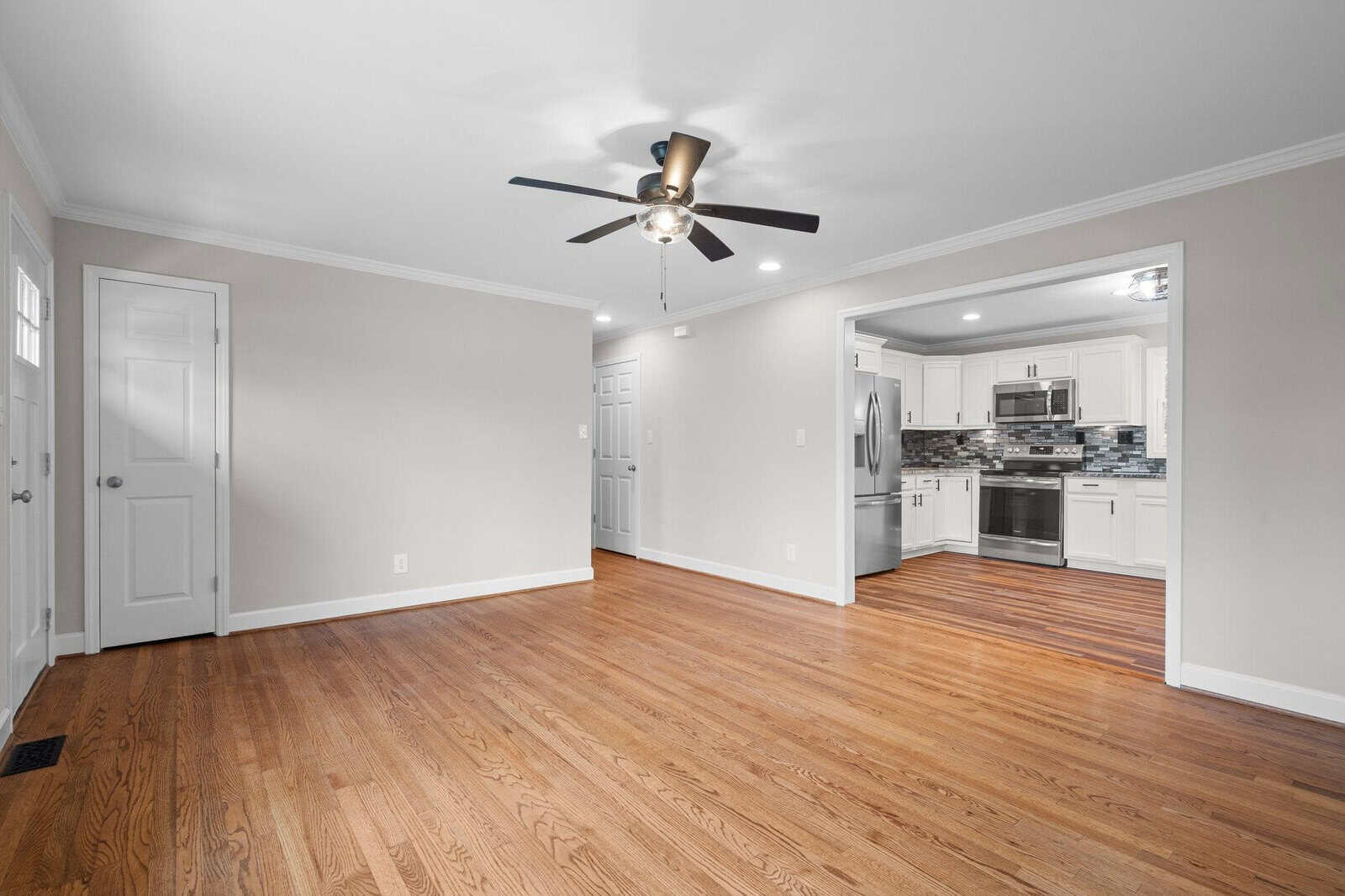 Modern, empty living space with hardwood flooring, white walls, and an overhead ceiling fan, leading to an adjoining kitchen with stainless steel appliances.