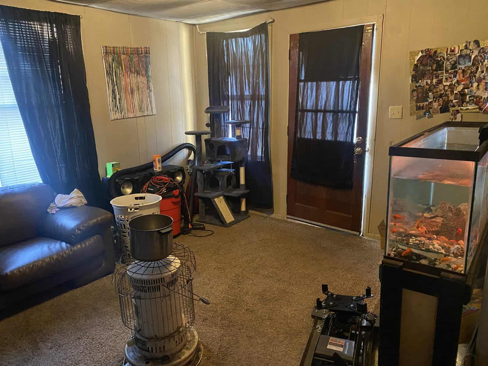 A living room with a variety of items including a blue sofa, an exercise bike, a cat tree, a kerosene heater, a fish tank, and walls decorated with curtains and numerous photos.