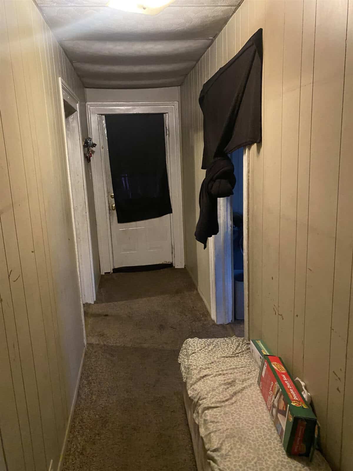 A narrow hallway with paneled walls and a carpeted floor, featuring a mattress on the right side, a hanging towel, and an open door at the end revealing a darkened room.