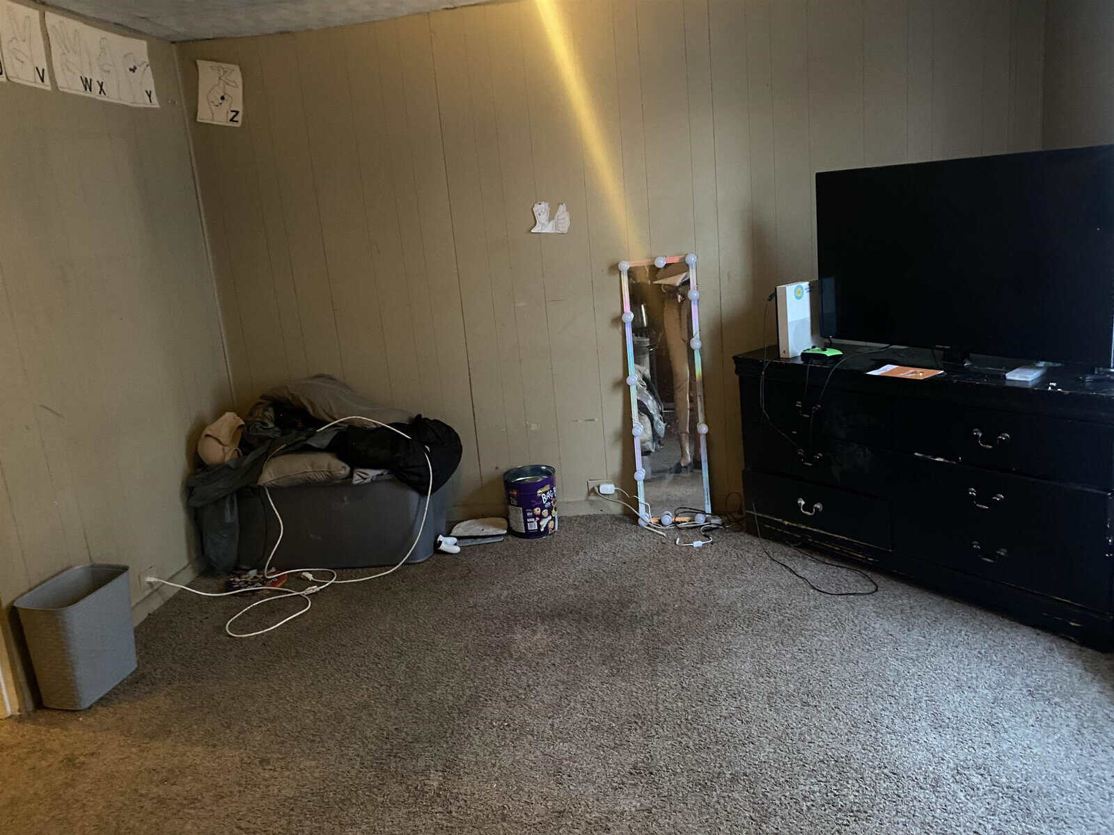 A room with beige walls and carpeted flooring containing a black television set on a dark cabinet, a pile of clothes on a chair to the left, a trash can, and assorted items strewn about.