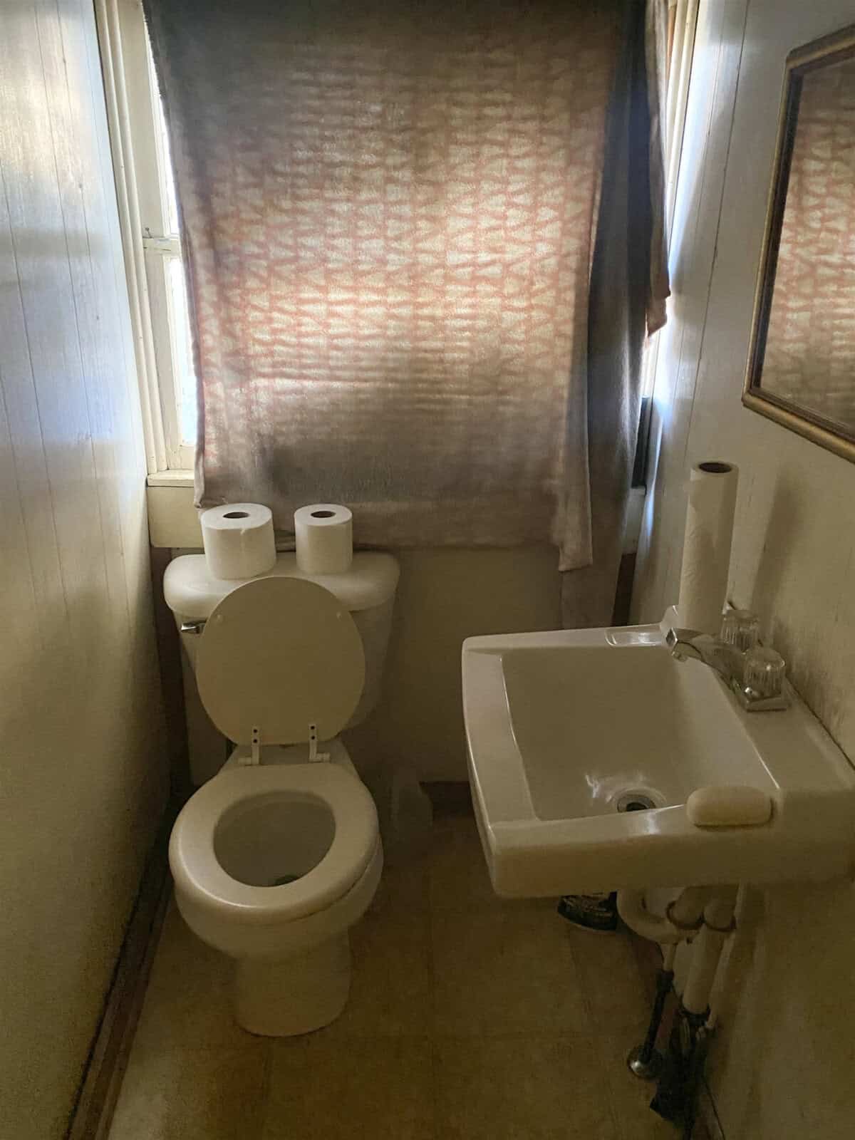A small, dimly lit bathroom with a toilet, a sink, and an opaque window covered by a patterned curtain, with a stack of toilet paper rolls on the toilet tank.