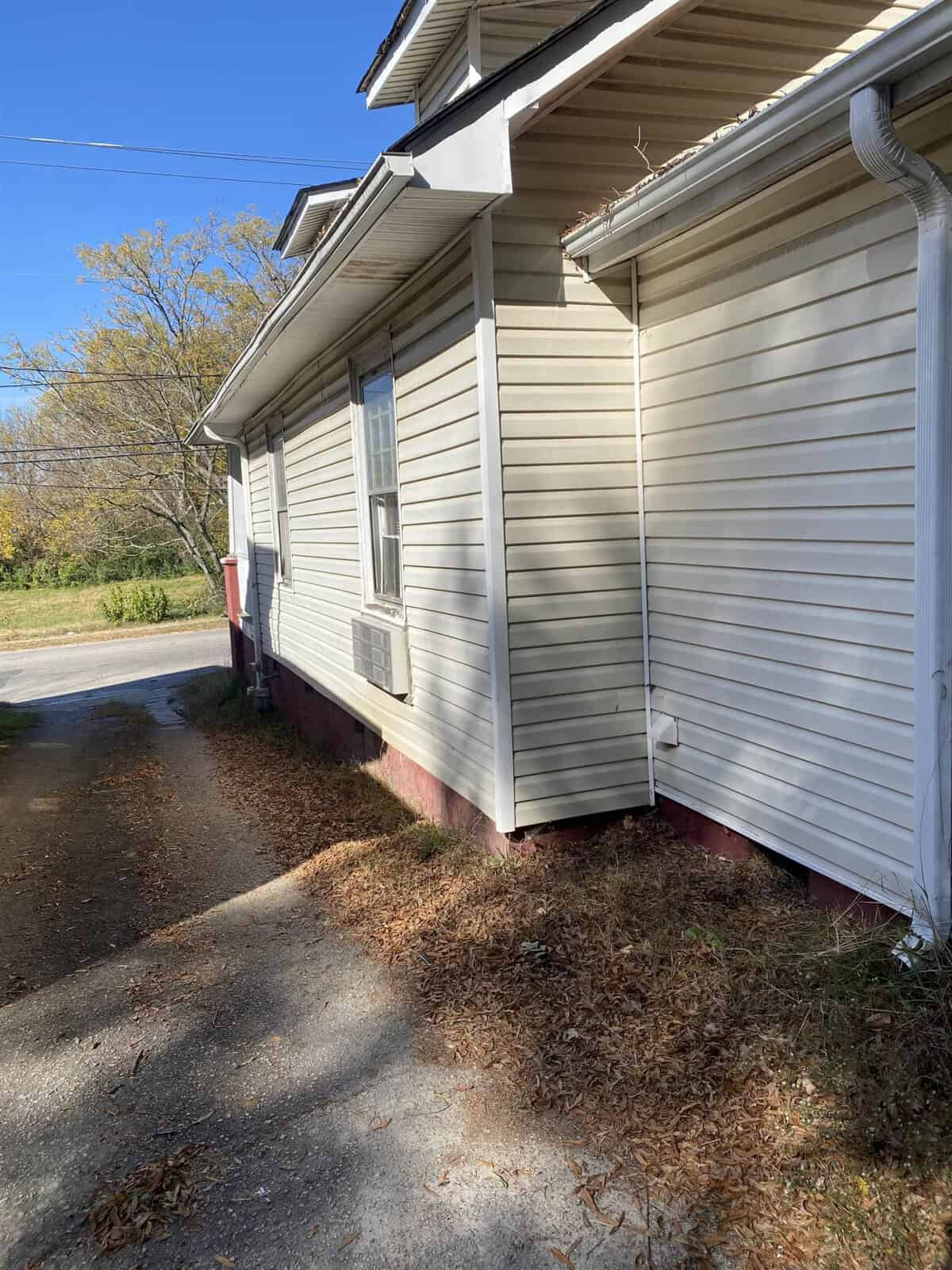 A suburban house with white siding and a red foundation viewed from the side, featuring a gutter downspout and a driveway with fallen leaves.