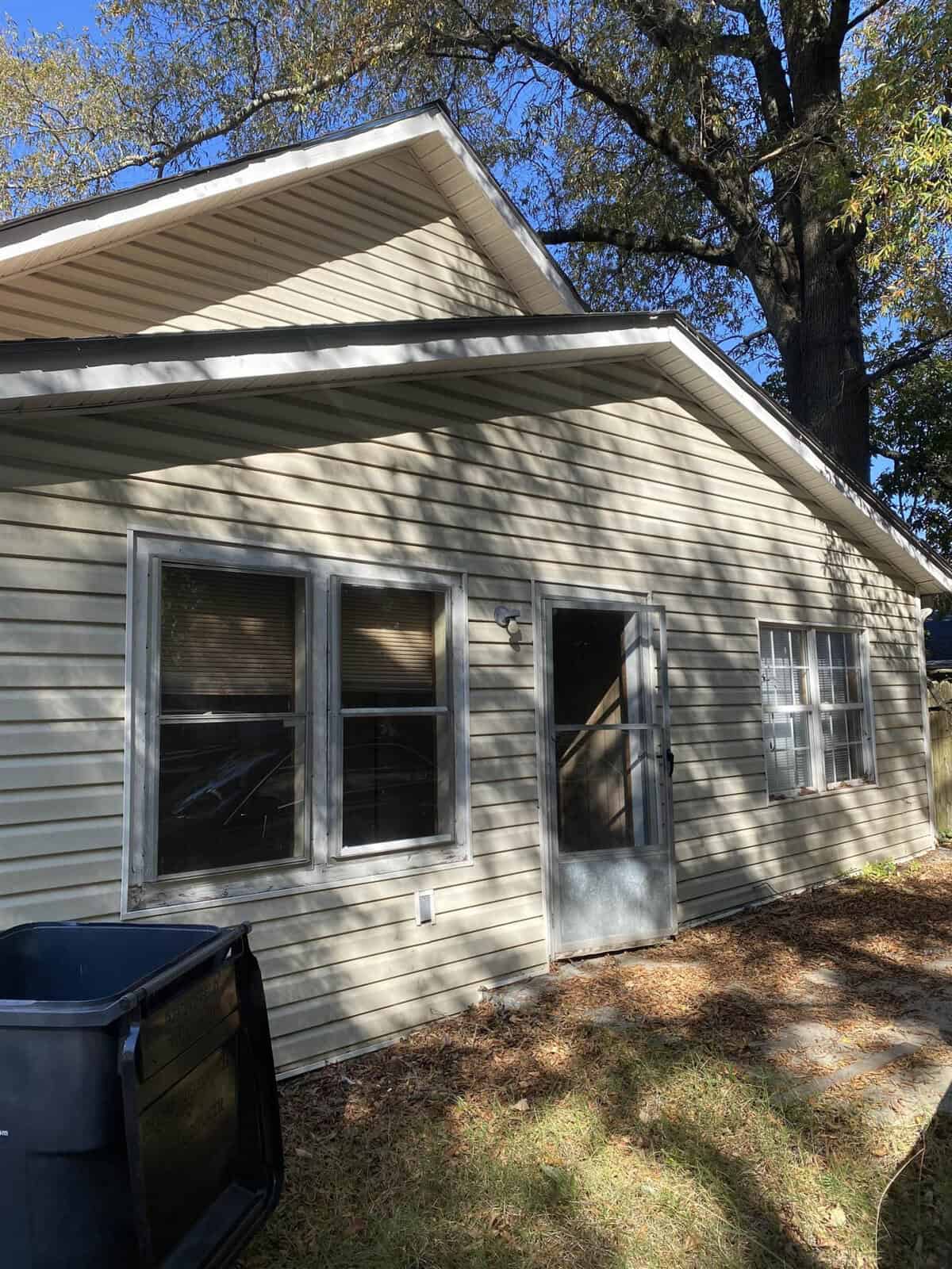 A single-story house with beige siding and white trim, featuring a set of windows and a glass paneled door, with a tree casting its shadow on the facade, and a trash bin to the left side on a concrete path.