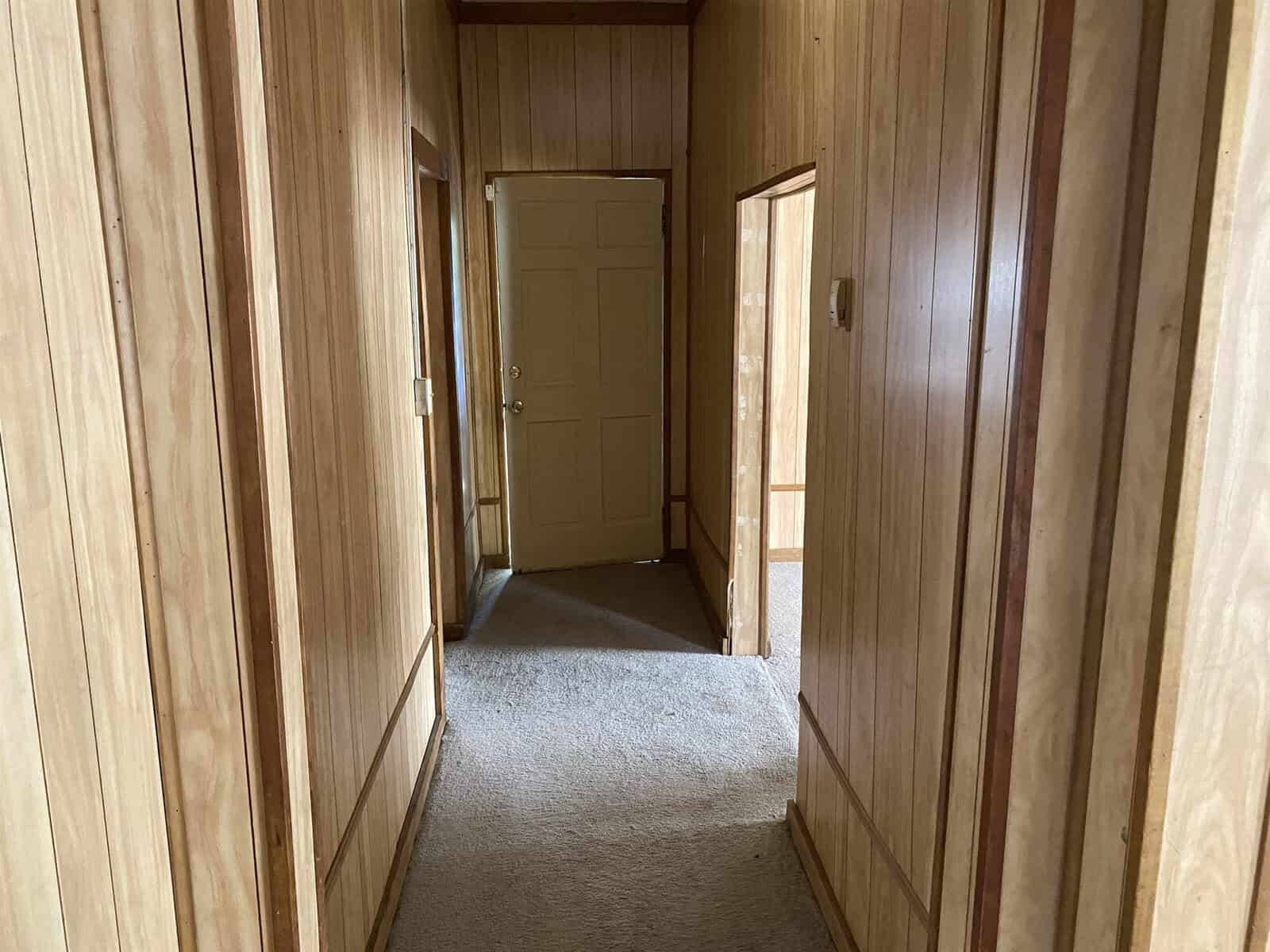 A narrow hallway in a house, lined with wooden paneling and beige carpeting, leading to a closed white door at the end.