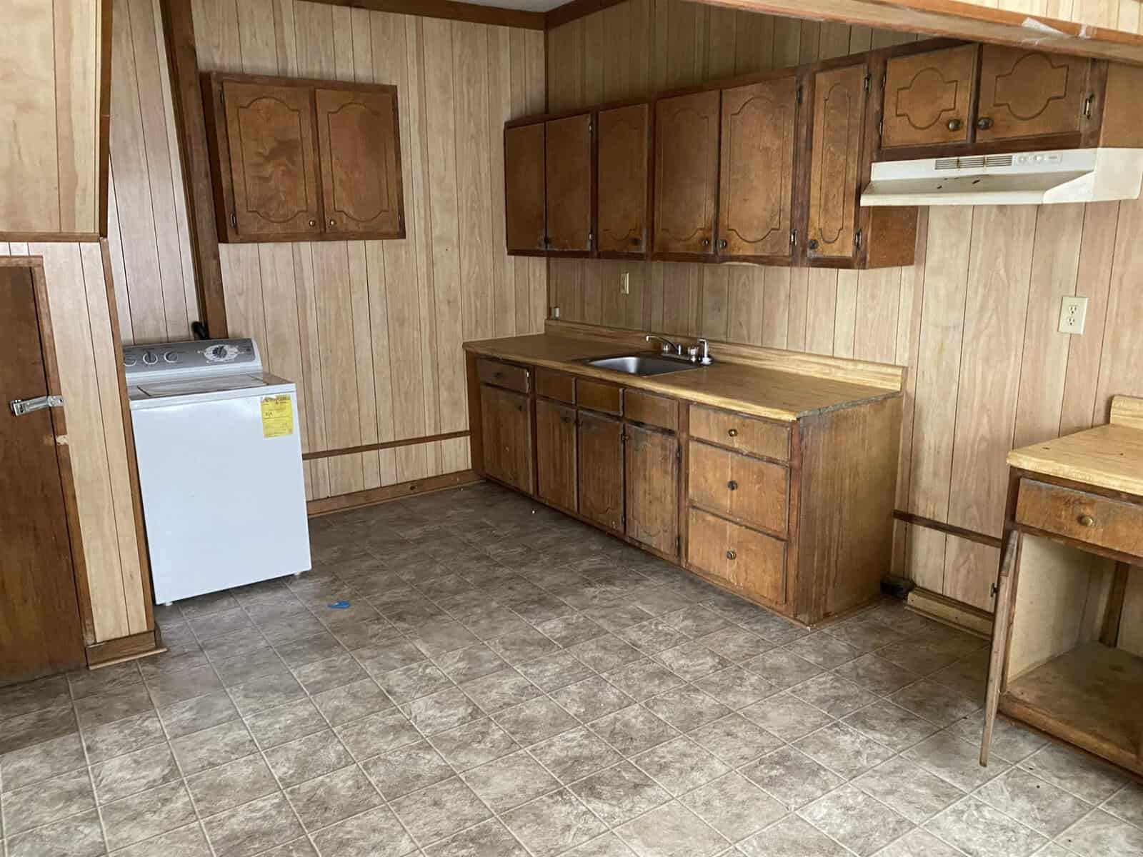 A dated kitchen interior with wood paneling, brown wooden cabinets, laminate countertops, a white refrigerator, and a sink with no visible appliances.