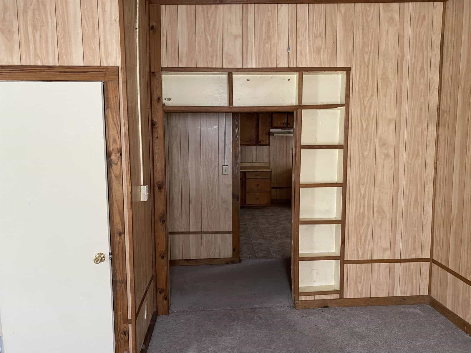 An empty room with wood paneling, featuring an open doorway leading to another room, and built-in shelving on the right side.