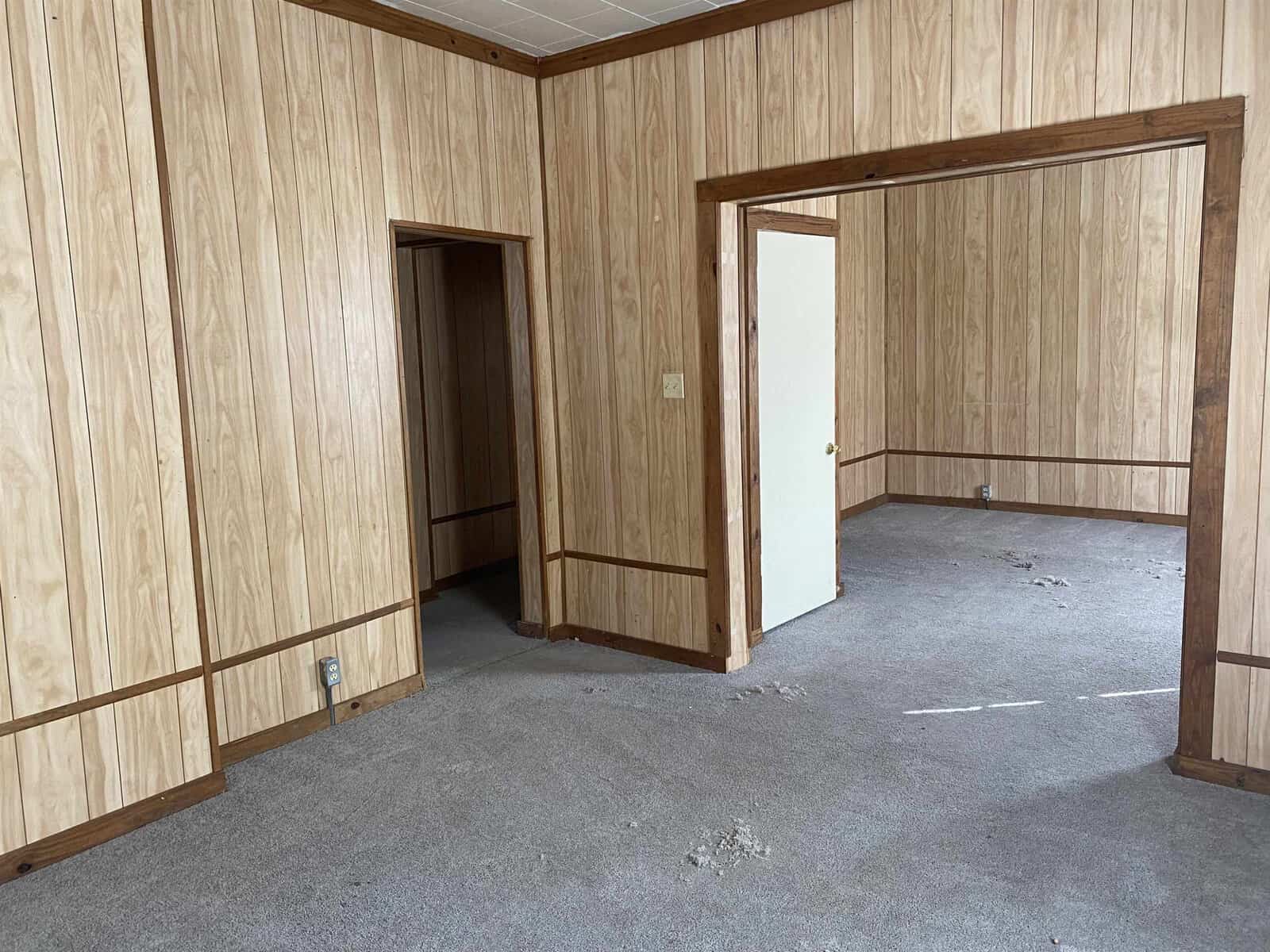 An empty room with wood paneling on the walls and a worn carpet on the floor. there is an open doorway to the left and a closed white door on the right.