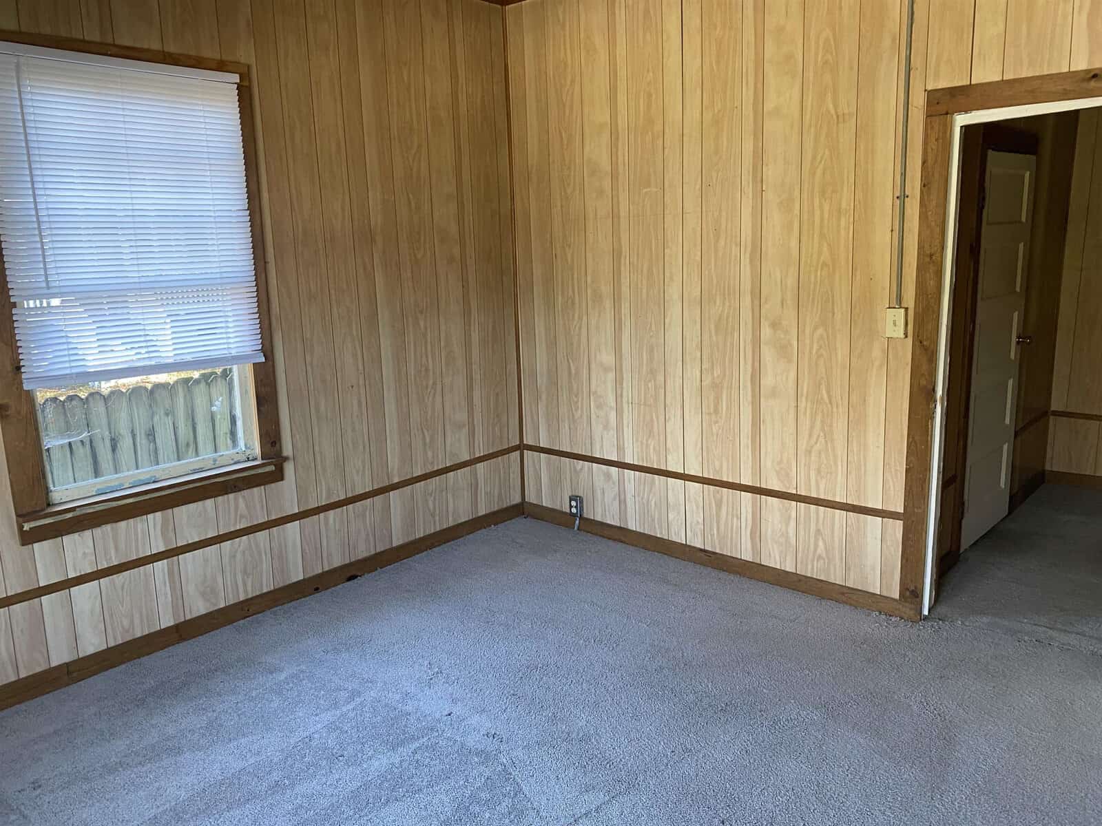 An empty room with wooden panel walls, a white window blind, beige carpeting, and a wooden baseboard. there's an open doorway on the right leading to another room.