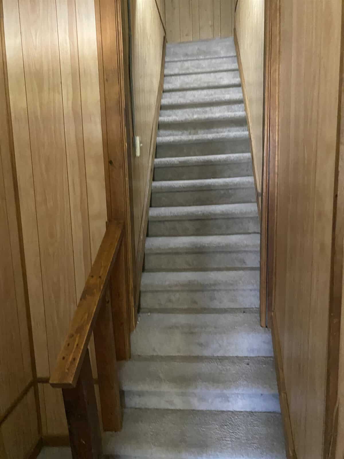 A narrow staircase with wooden panel walls and a wooden railing on the left, leading upwards between the walls. the steps appear worn, indicating frequent use over time.