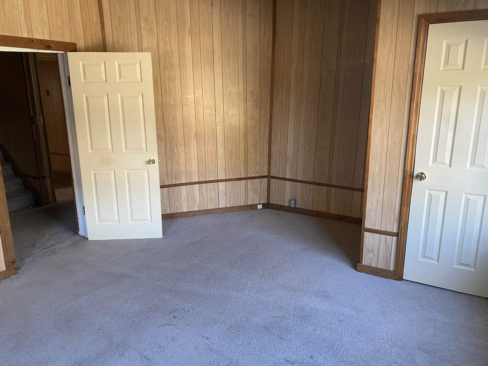 An empty room with wood paneling, two closed white doors, and beige carpeting.