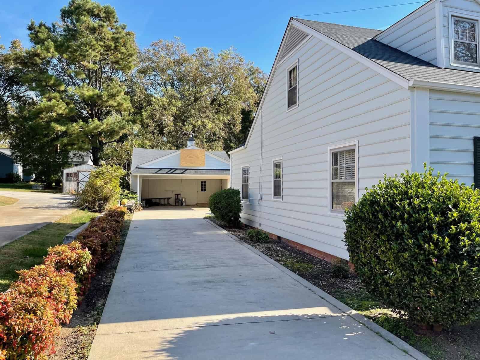 A well-maintained footpath leads to a detached garage beside a two-story residential home, flanked by manicured hedges and trees under a clear blue sky.