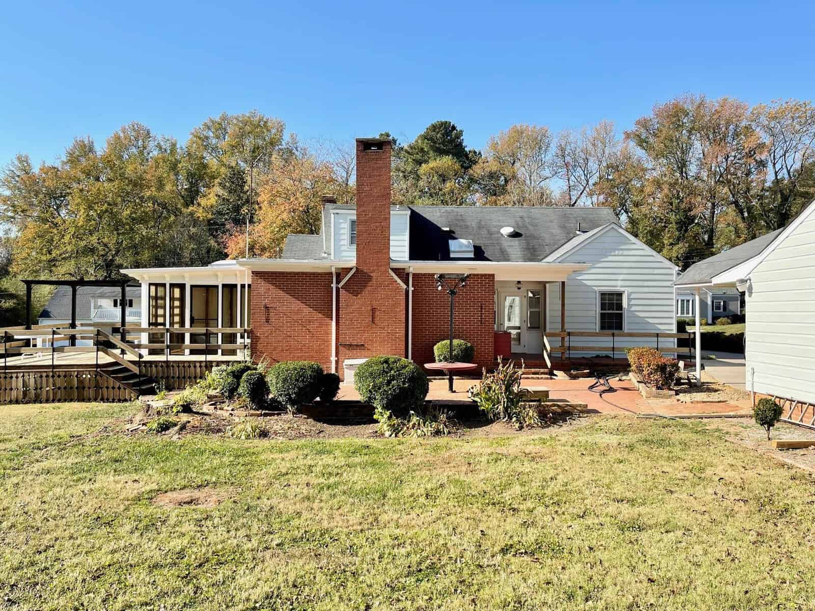 A single-story brick house with a white extension, featuring a chimney and a wooden deck area in a suburban setting.
