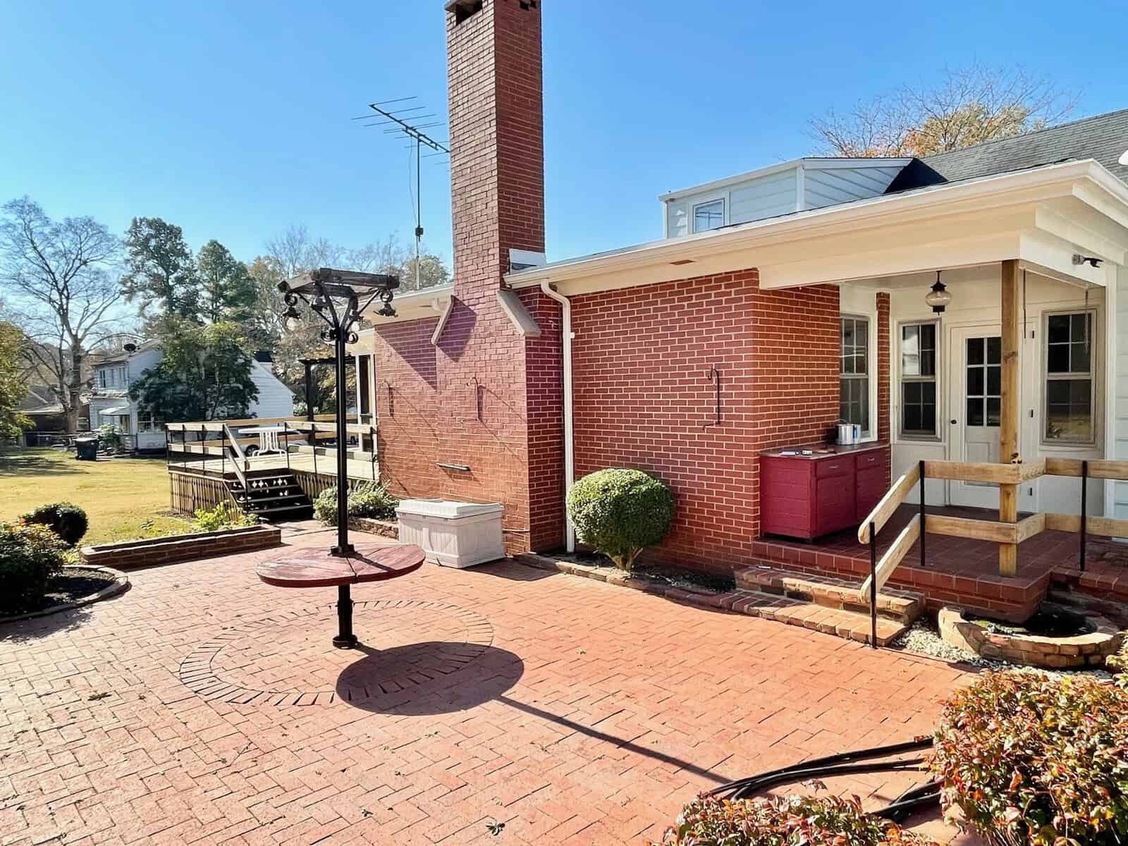 A brick house with a white porch and wooden stairs overlooking a paved patio with a circular design and a metal bird feeding station.