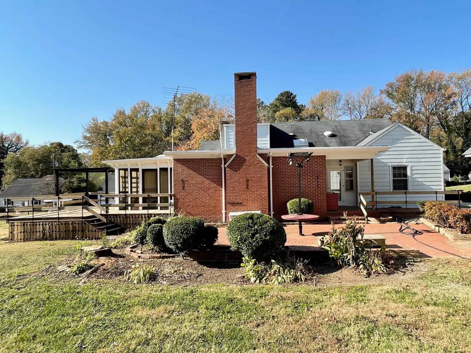 A single-story brick house with a white extension and chimney, featuring a spacious deck, surrounded by a grassy lawn with shrubs, under a clear blue sky.