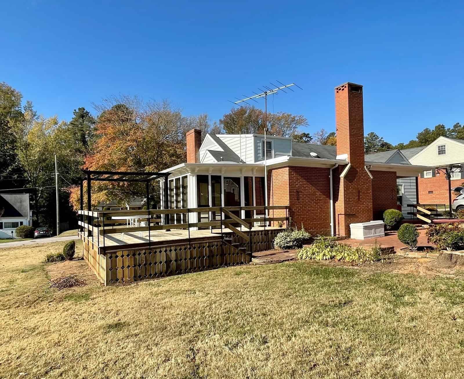 A single-story brick house with a white porch, an attached wooden deck, and a prominent chimney, set against a background of trees with autumn foliage.