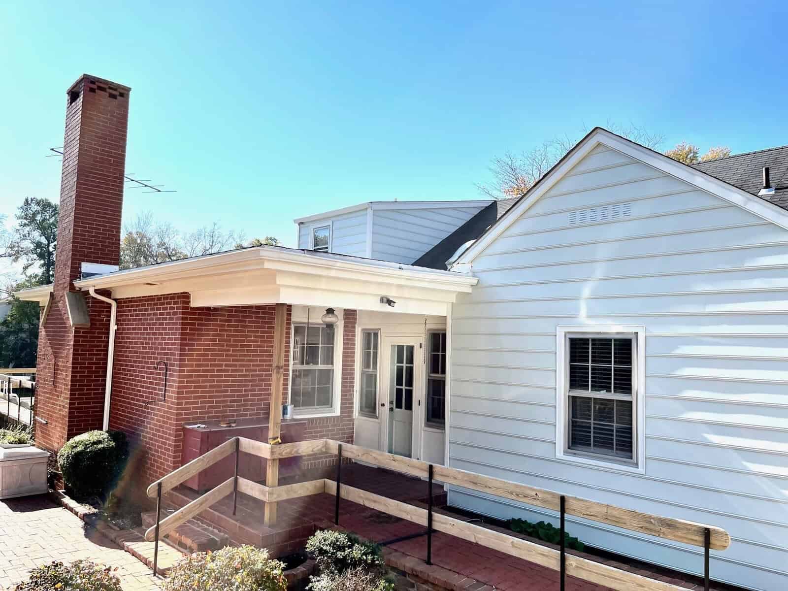 A sunny view of a single-story white house with a brick chimney and steps leading to a back entrance.