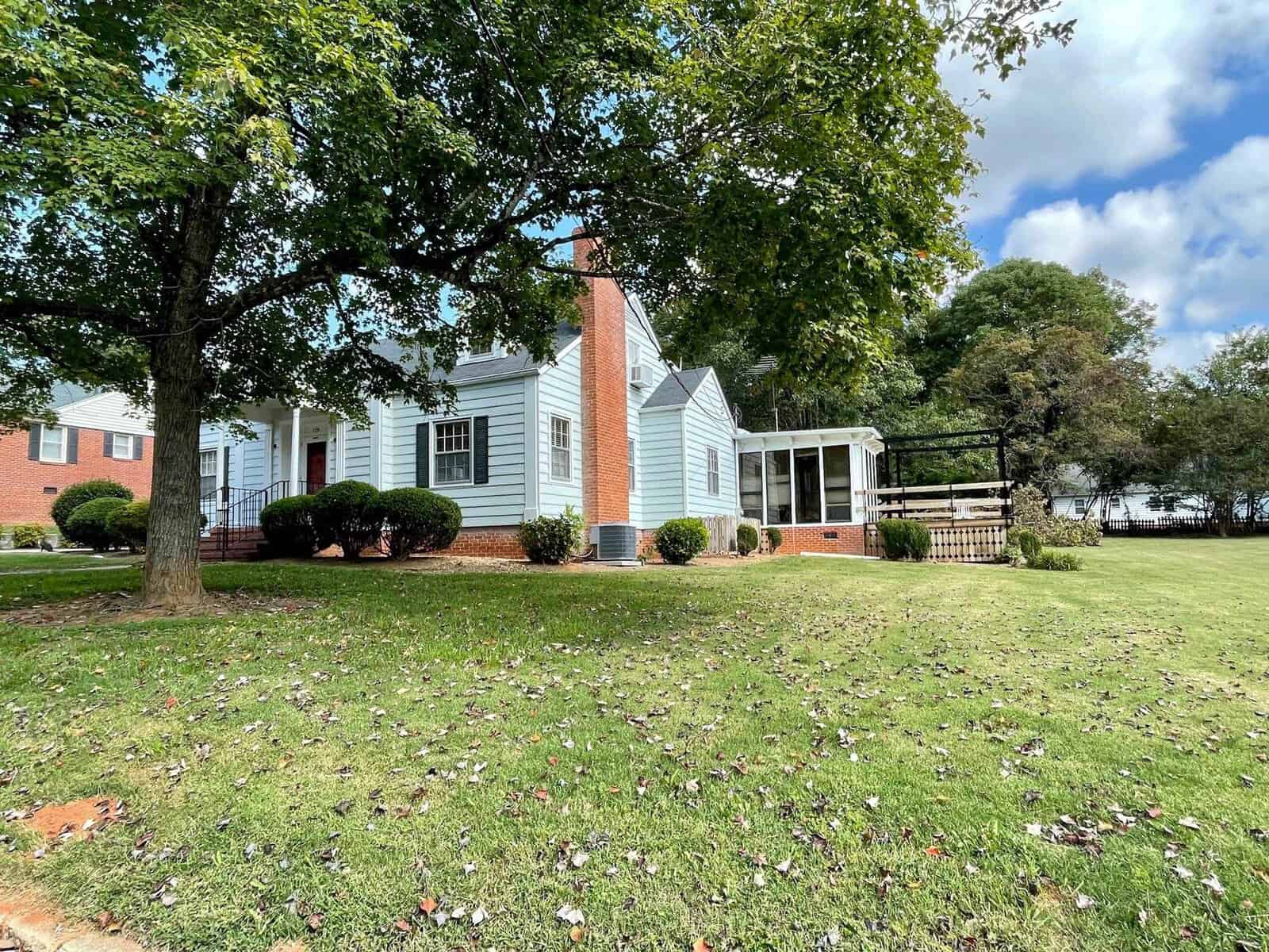 A single-story white house with a brick chimney and porch, surrounded by a lawn with scattered leaves under a partly cloudy sky.
